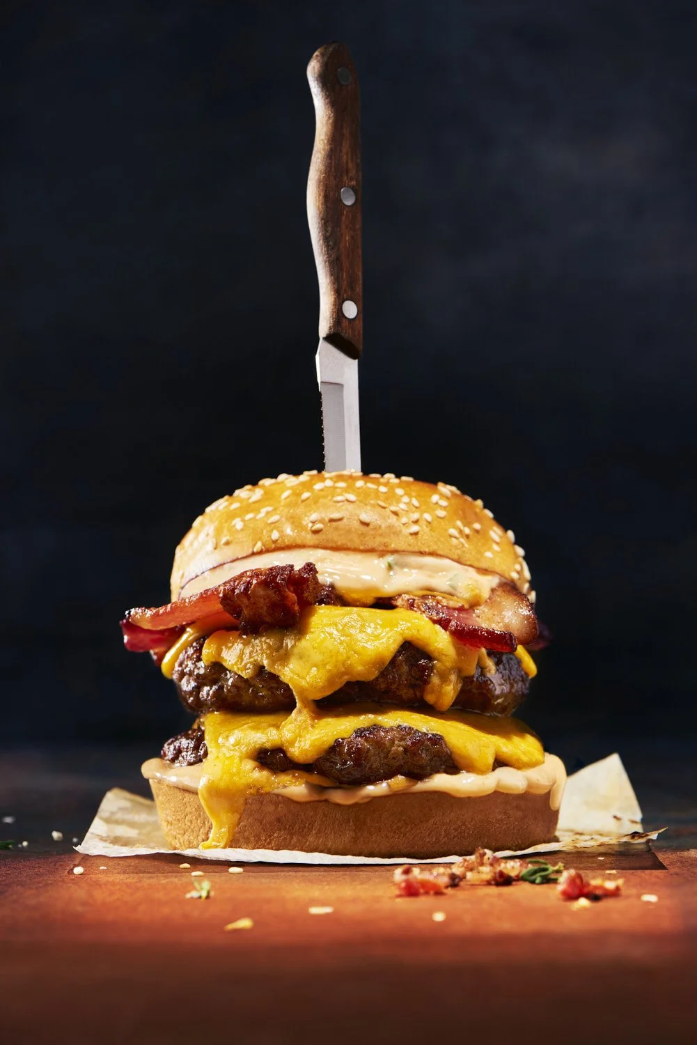 Dark dramatic close-up of a towering double cheeseburger with a knife plunged through the center, moody low-key studio lighting against a near-black background