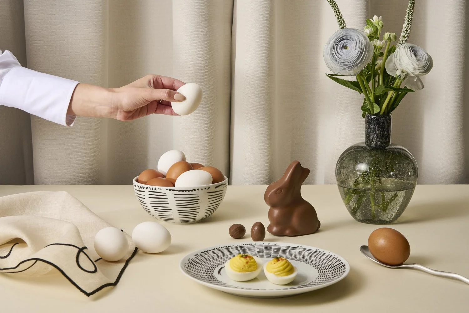 Elegant Easter or spring table setting with a ceramic bunny, eggs, nuts, and florals on a neutral linen surface, soft natural light with warm styling