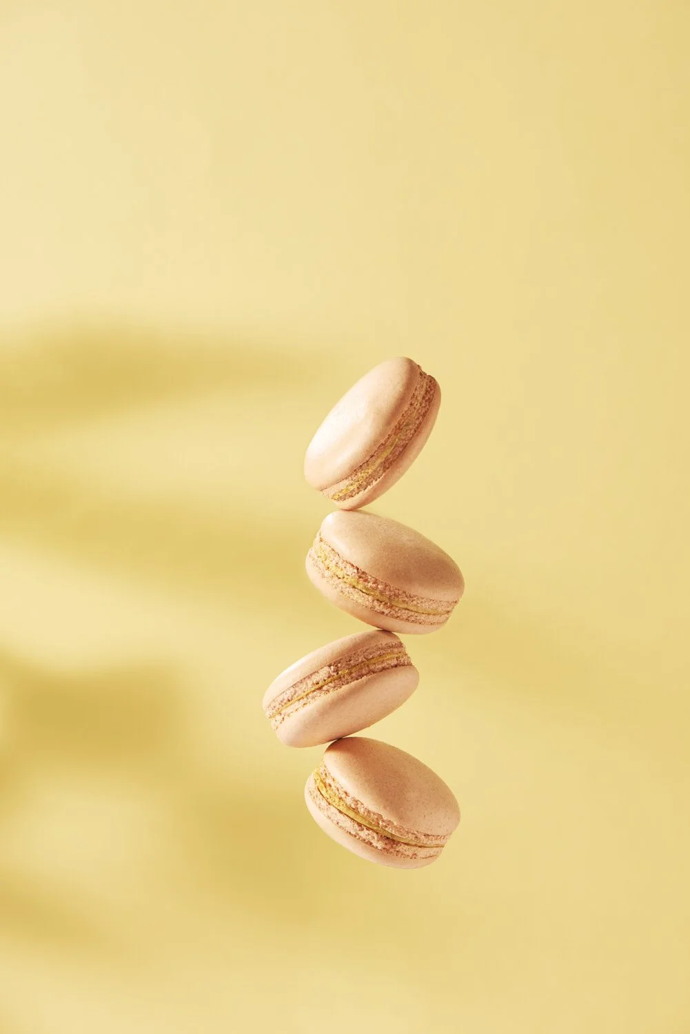 Four stacked macaroons resting on a warm golden yellow background, lit softly from one side to highlight texture and colour