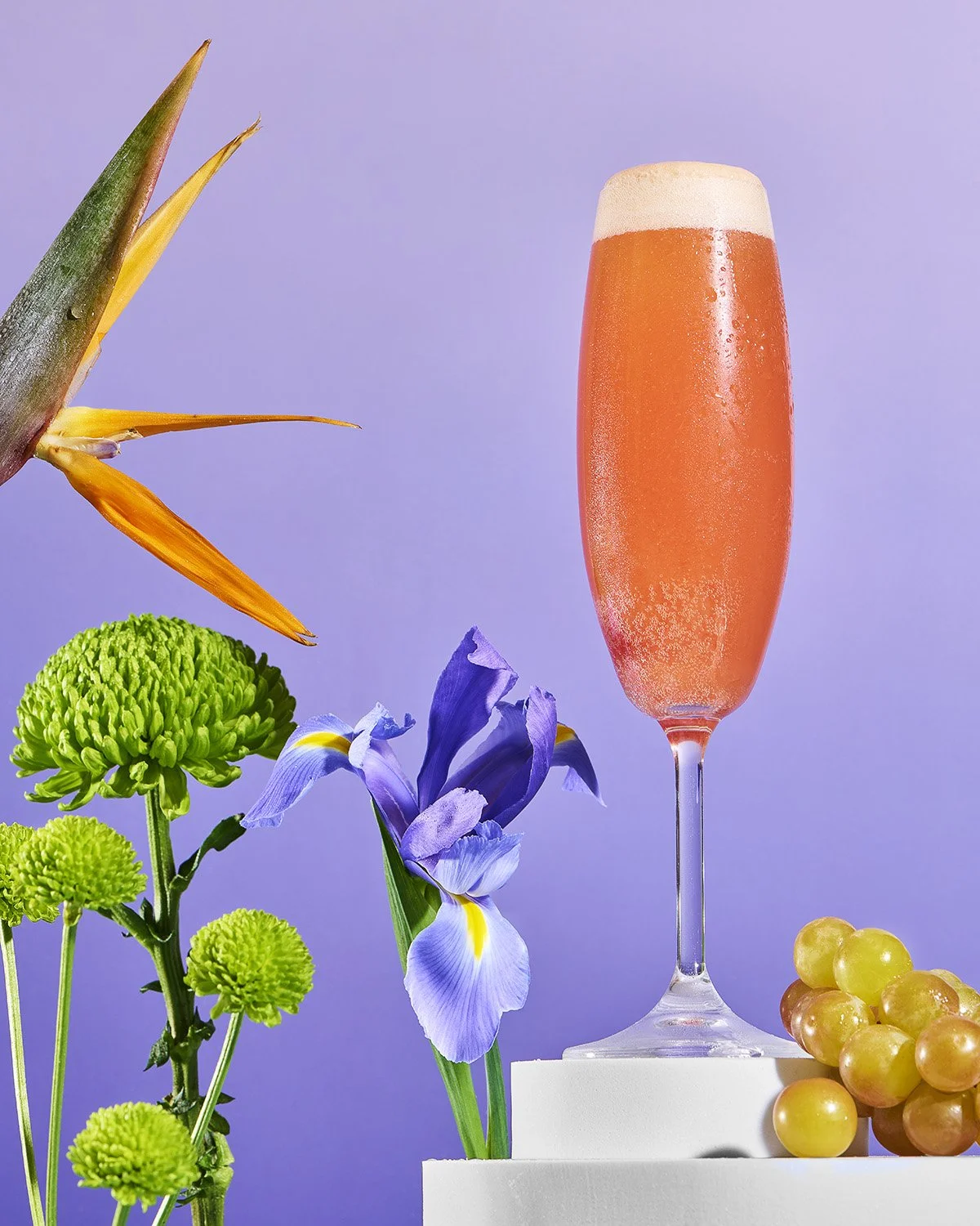 Tall champagne flute filled with a pink Bellini or rosé cocktail, styled with tropical bird of paradise flowers and purple irises on a lavender background