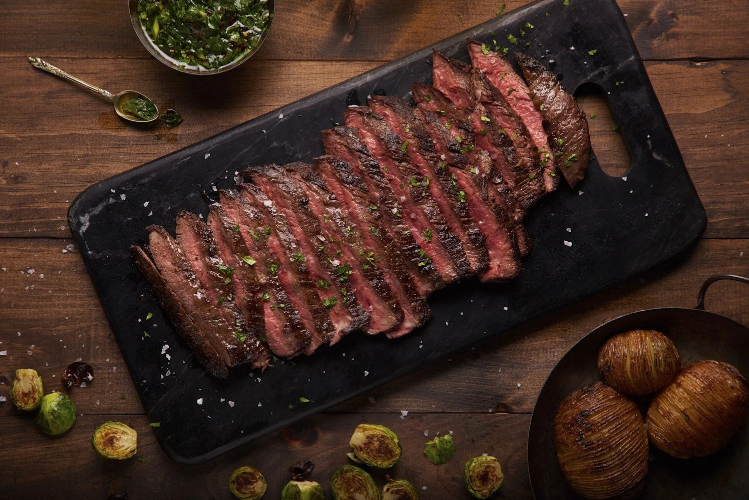 Thinly sliced grilled flank steak fanned out on a dark slate board, garnished with fresh herbs and peppercorns, shot with dramatic side lighting from above
