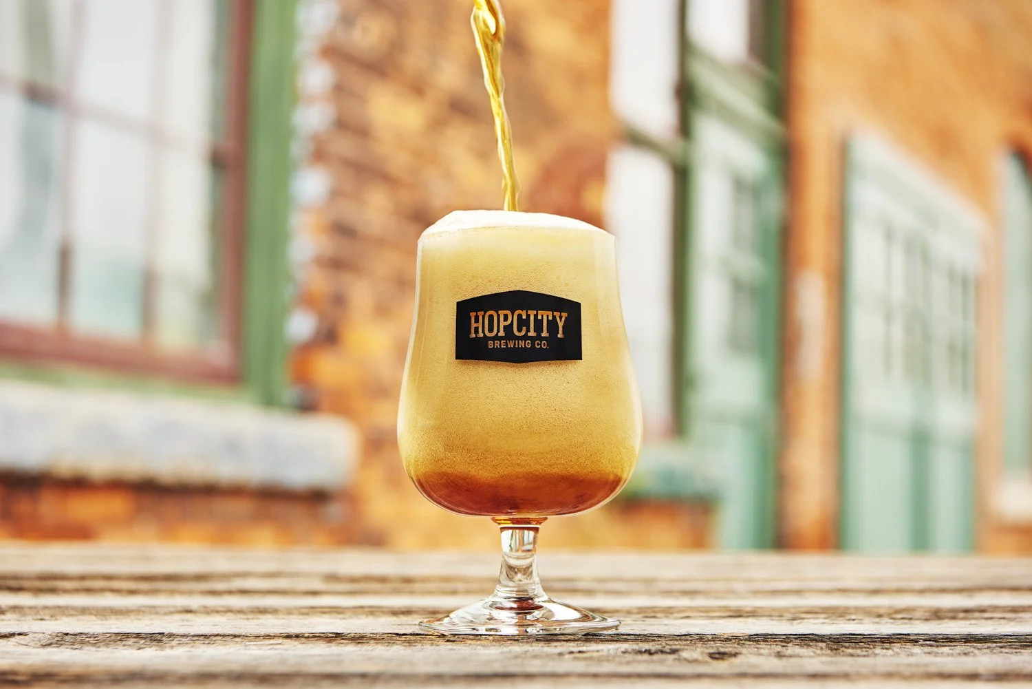 Branded cocktail in a tall glass served on a wooden outdoor patio table, warm natural sunlight with a colorful urban building facade in the background