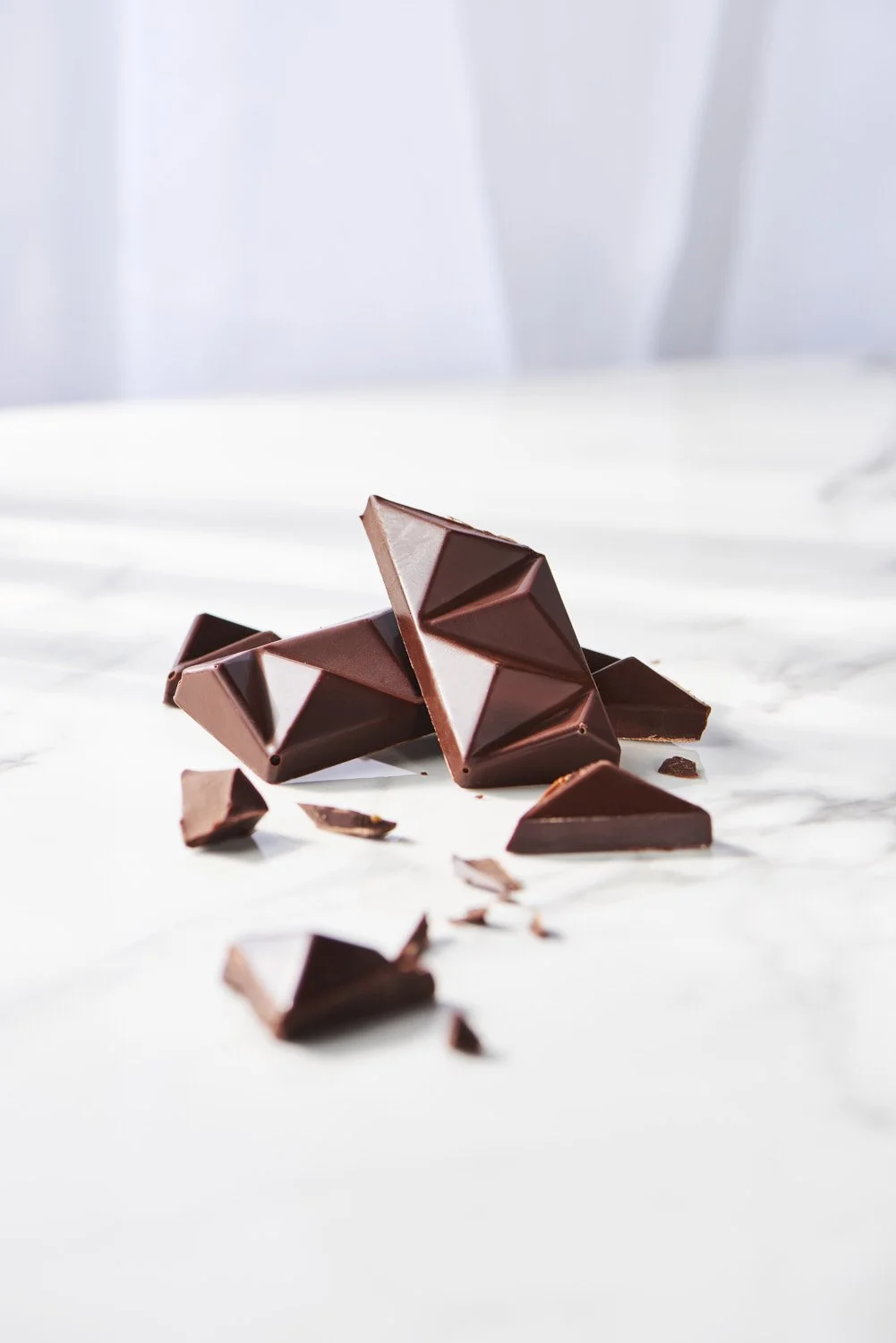 Scattered broken dark chocolate pieces and shards arranged on a clean white surface, minimal flat lay product shot with soft natural light and clean shadows