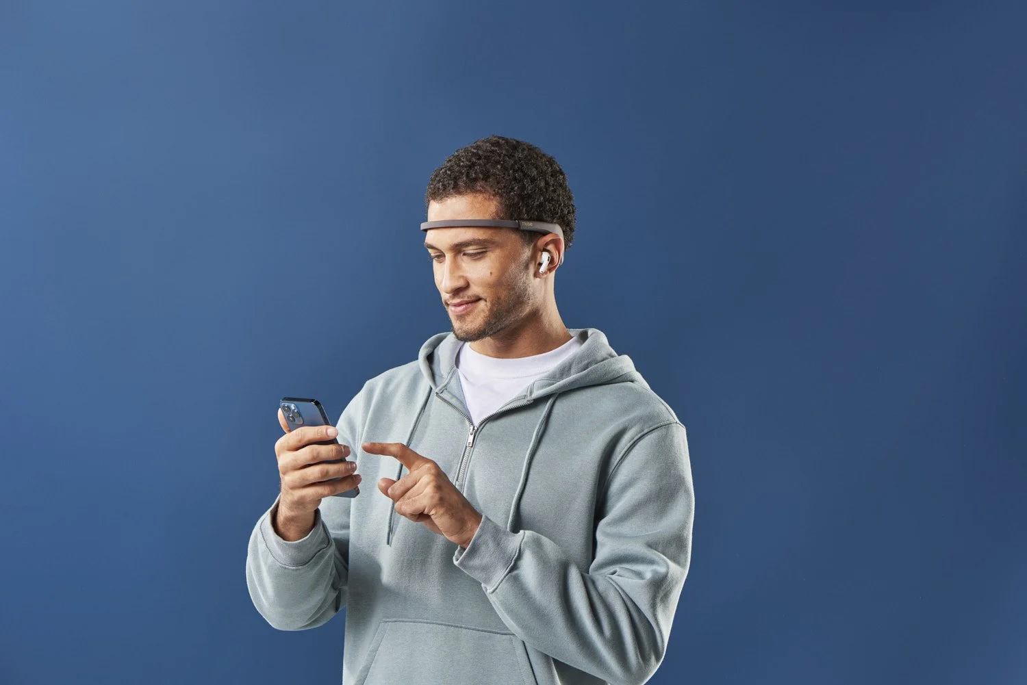 Young man in a grey hoodie holding a small cup while wearing a tech headband wearable, casual lifestyle portrait against a solid dark blue background