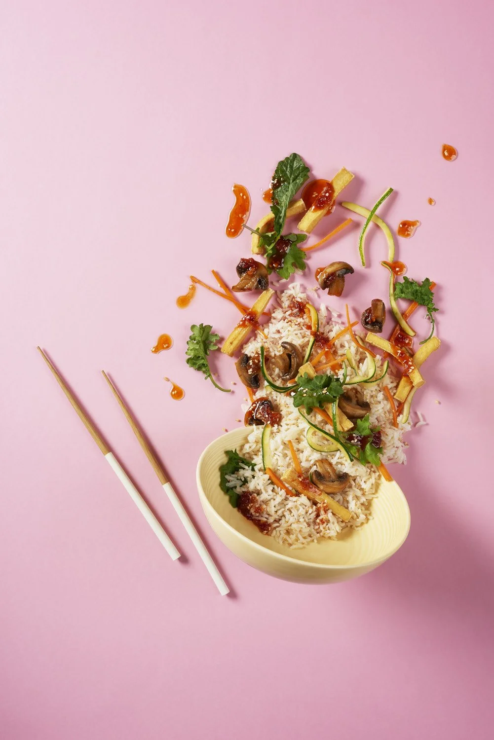 Fresh rice bowl scattered with vibrant herbs, edible flowers, and thinly sliced vegetables, photographed flat lay on a clean pastel pink background with soft light