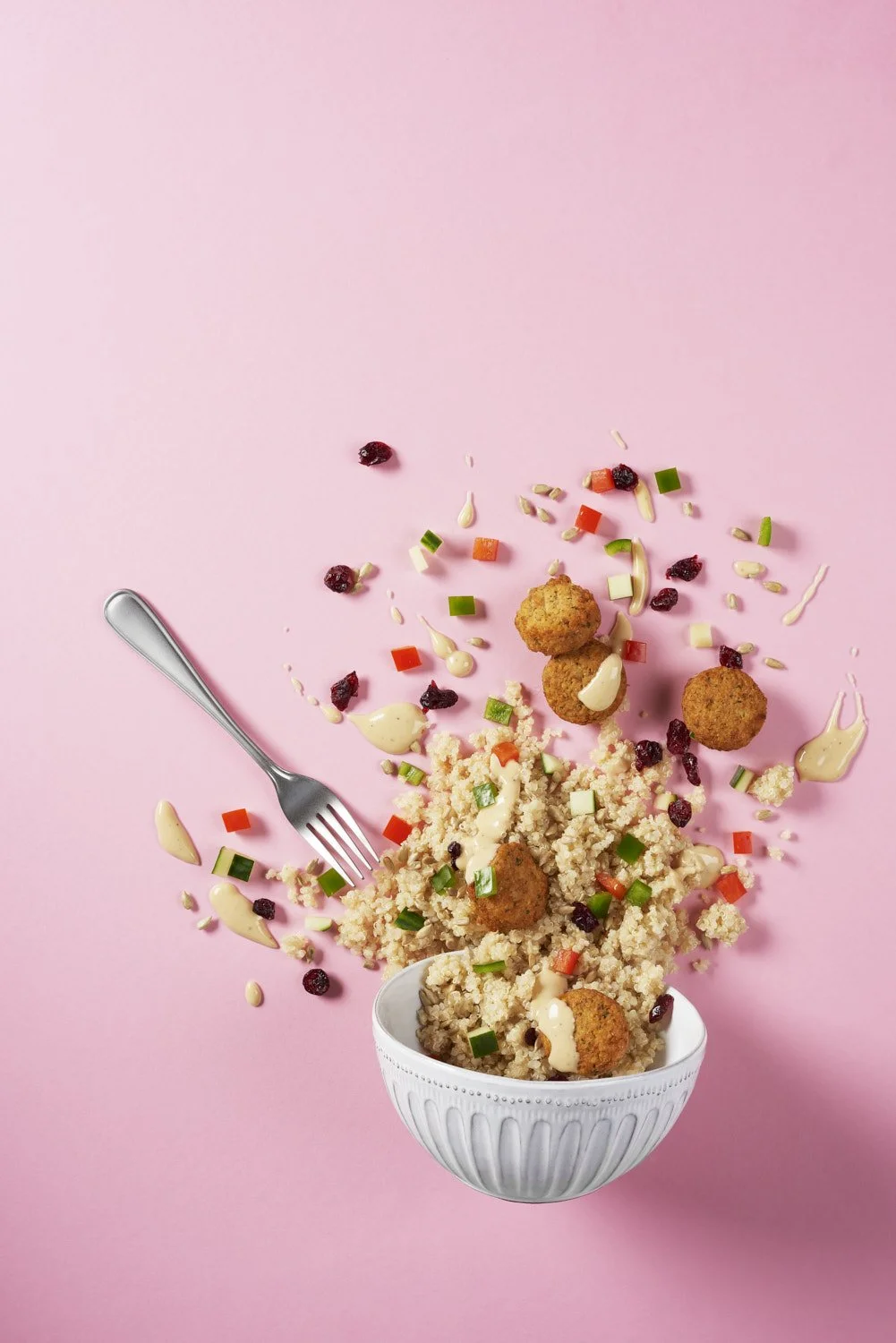  Artistic exploding grain bowl with seeds, dried flowers, herbs, and toppings suspended mid-air above a white bowl, shot on a clean pastel pink background