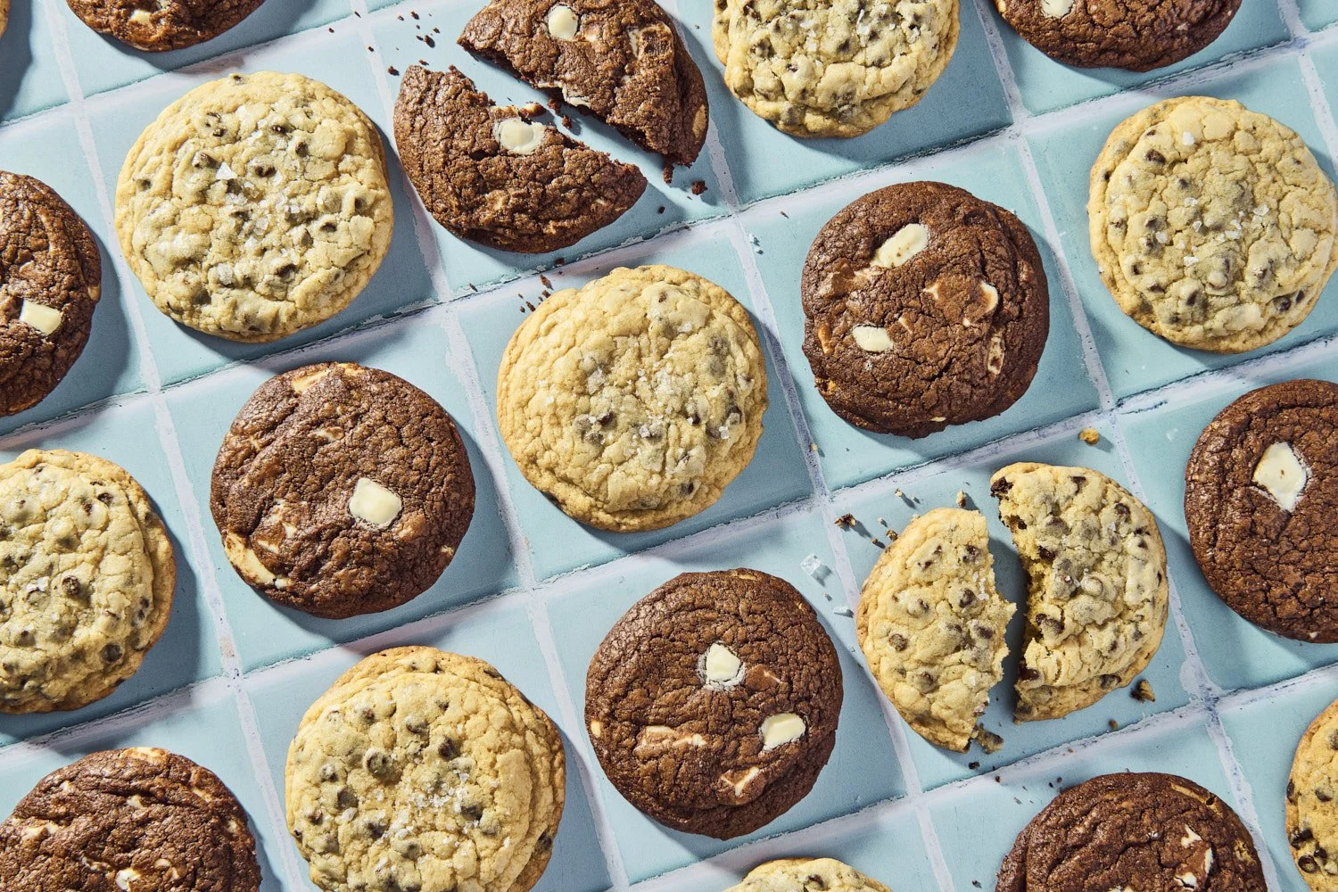 Assorted chocolate chip and frosted sandwich cookies arranged in a grid pattern on a light blue baking tray, shot from directly overhead on a matching surface