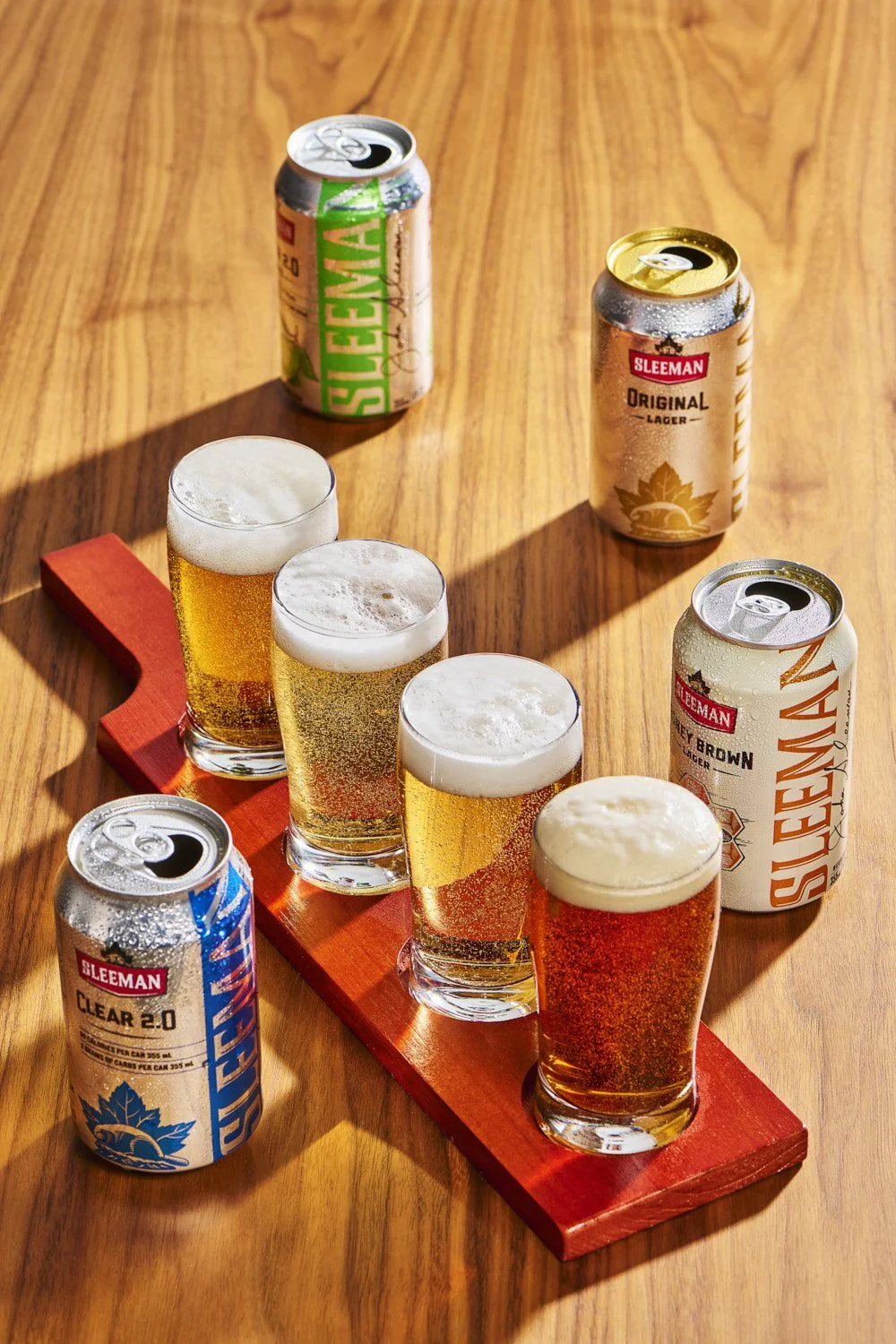 Multiple branded canned beverages displayed upright on a wooden serving board with small glasses, colorful product lineup shot in warm natural indoor light