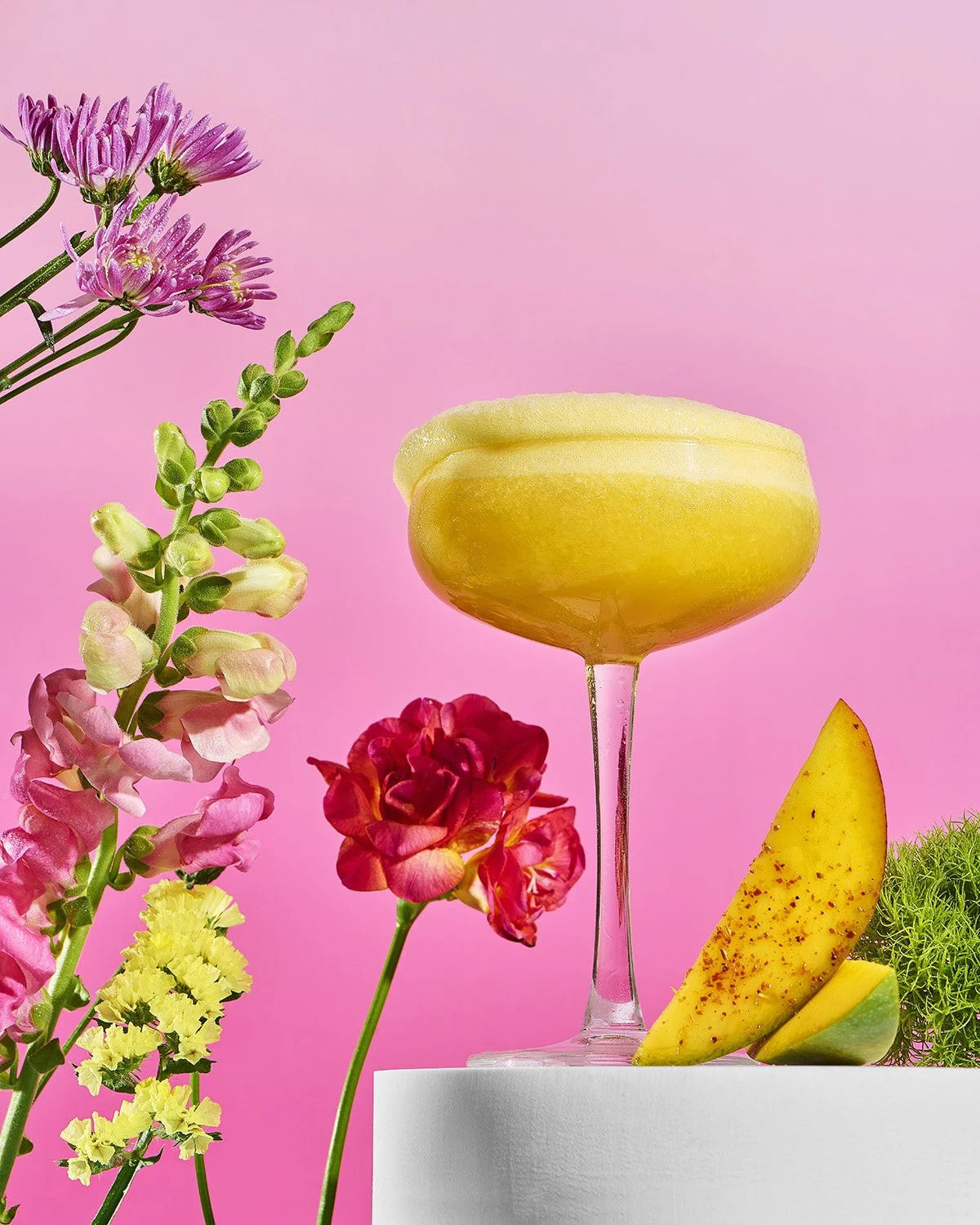 Bright yellow margarita-style cocktail in a coupe glass surrounded by tropical flowers and a slice of mango on a vibrant hot pink background