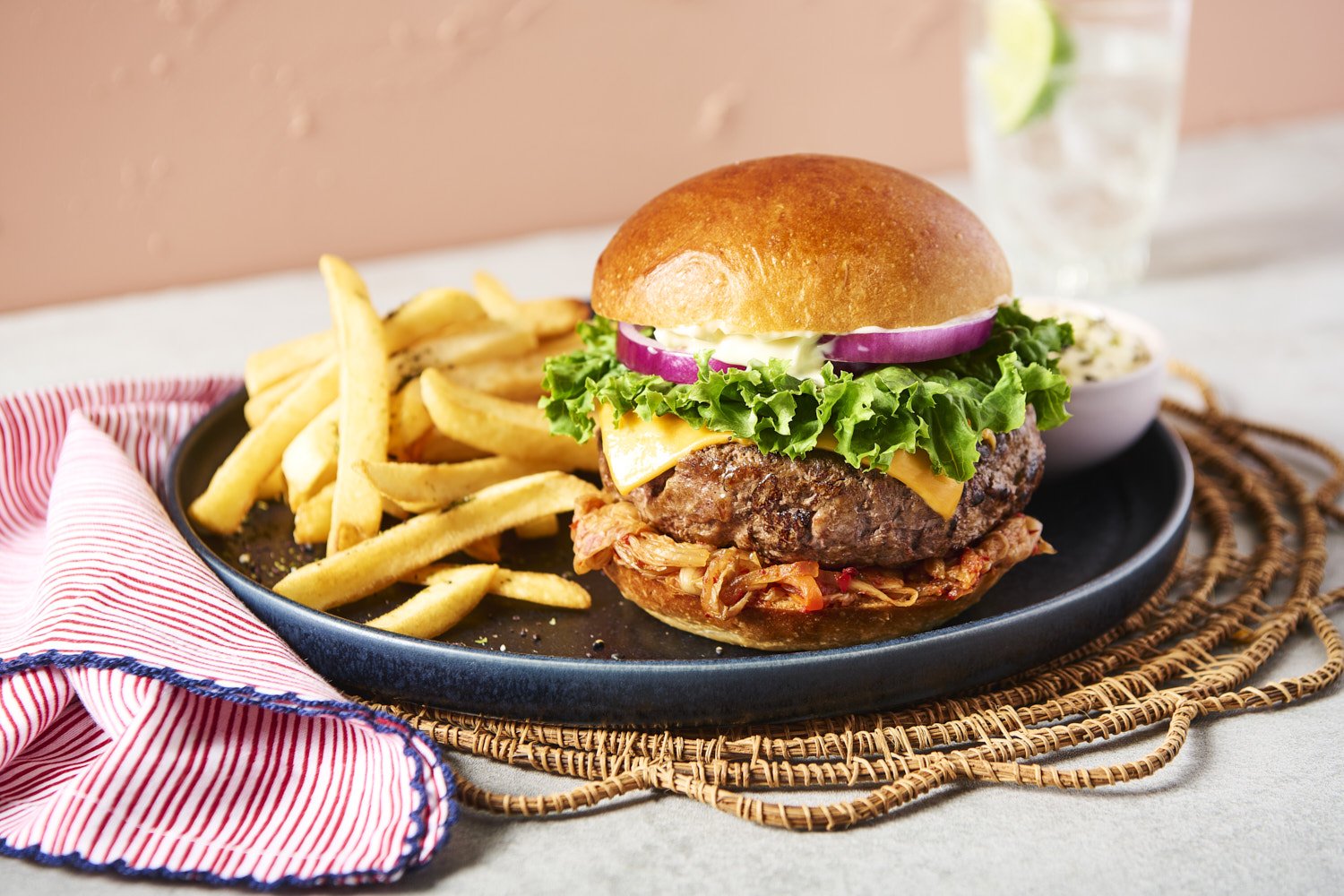 Burger withered onion,  slaw and sauce on a dark wicker placemat, with a side of seasoned french fries and a drink