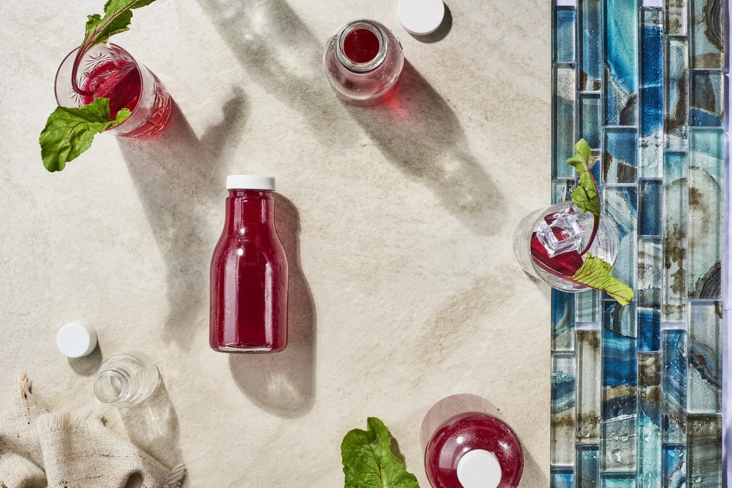 Overhead flat lay of a dark berry juice bottle and fresh cranberries scattered on a teal blue mosaic tile surface with soft even lighting