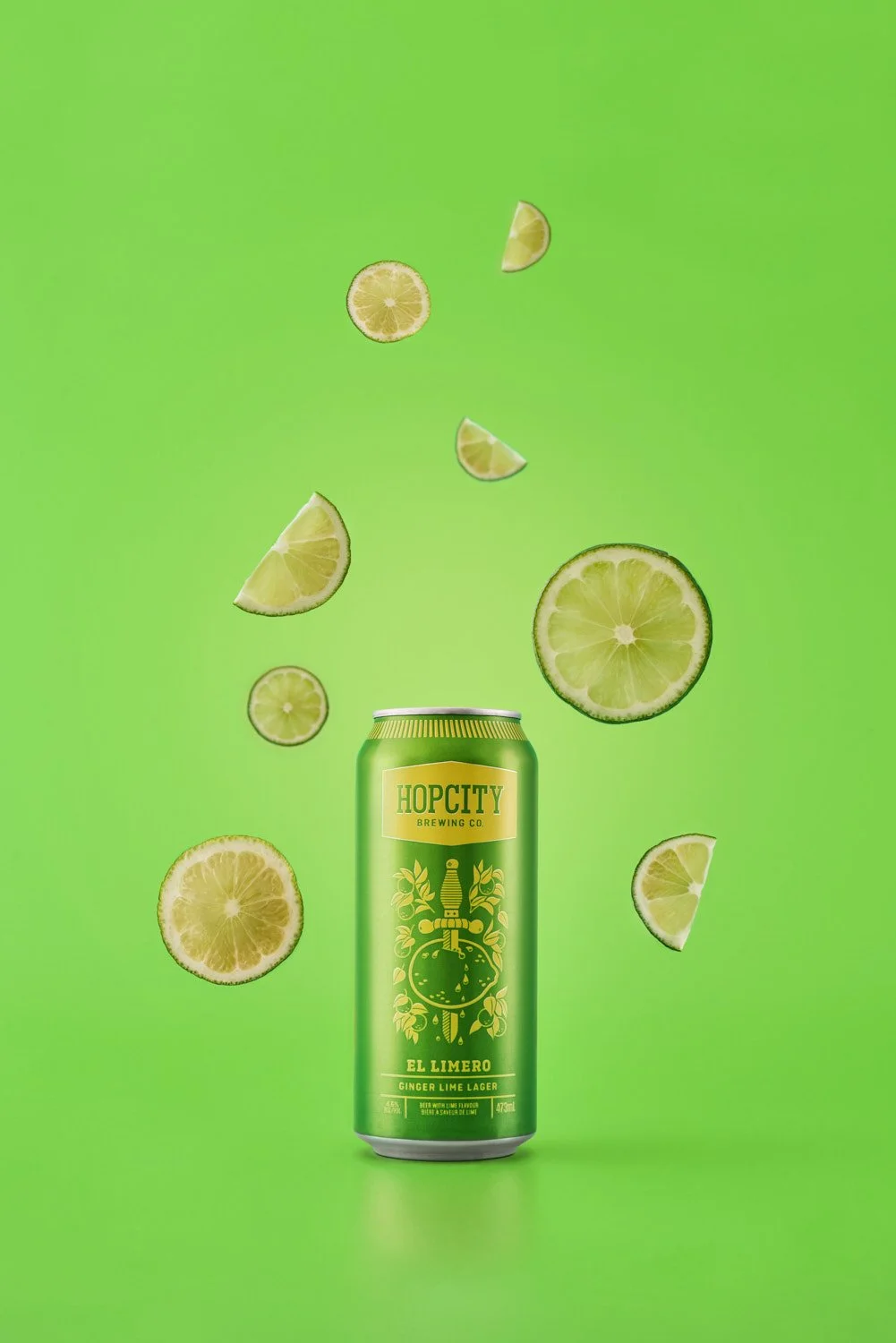 Branded beer can on a vivid green background with lime slices and citrus pieces falling around it, energetic product shot with bold color contrast