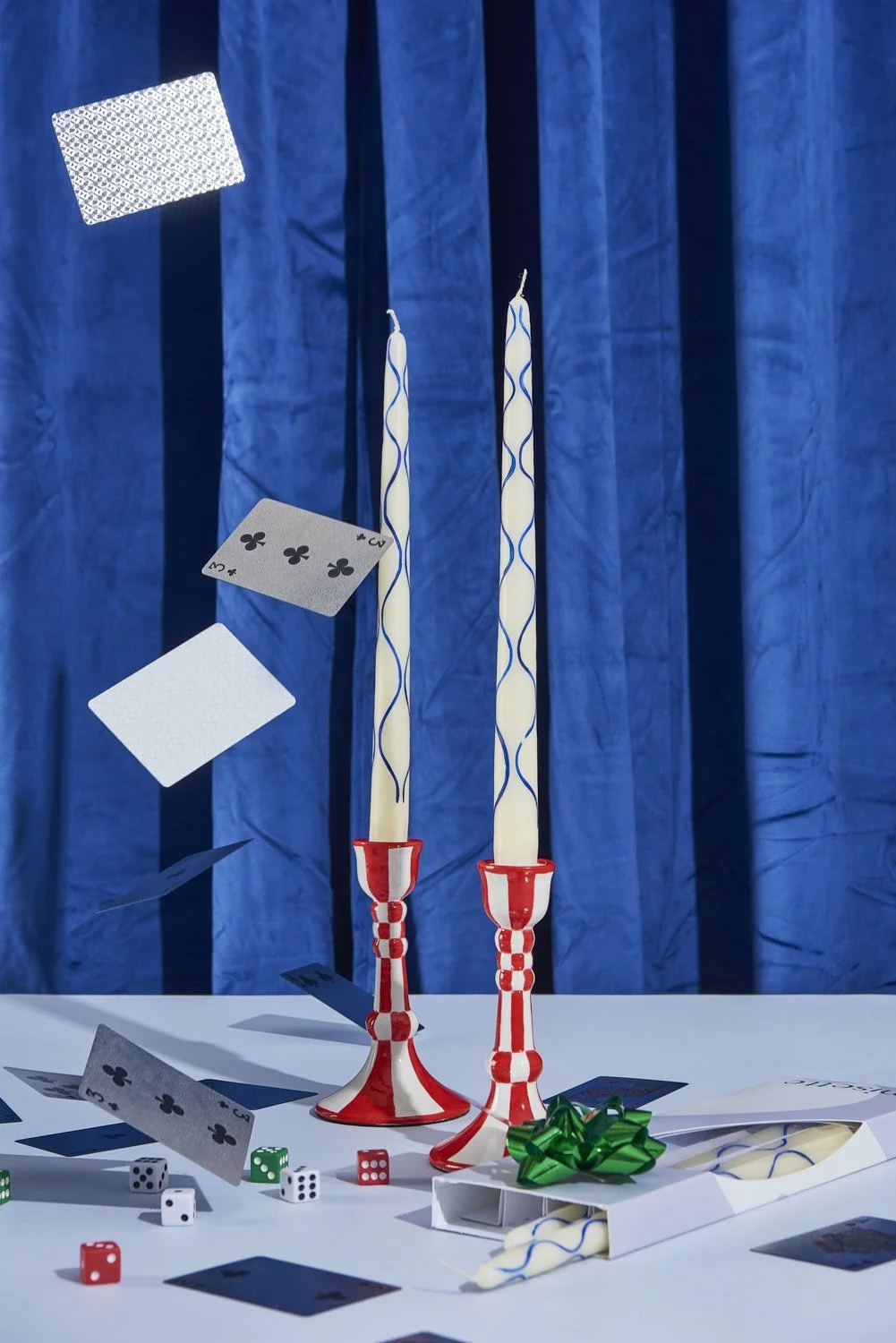 Artistic still life with tall taper candles, small stacked boxes, and decorative objects arranged against a deep royal blue velvet curtain background with dramatic lighting