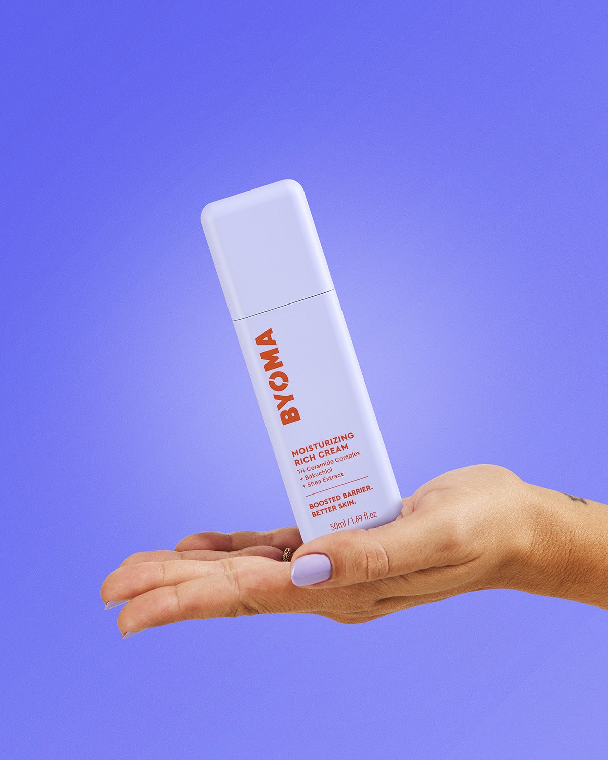 Hand holding a white Byoma skincare tube against a vibrant blue to purple gradient background, clean product-in-hand lifestyle shot with smooth even lighting