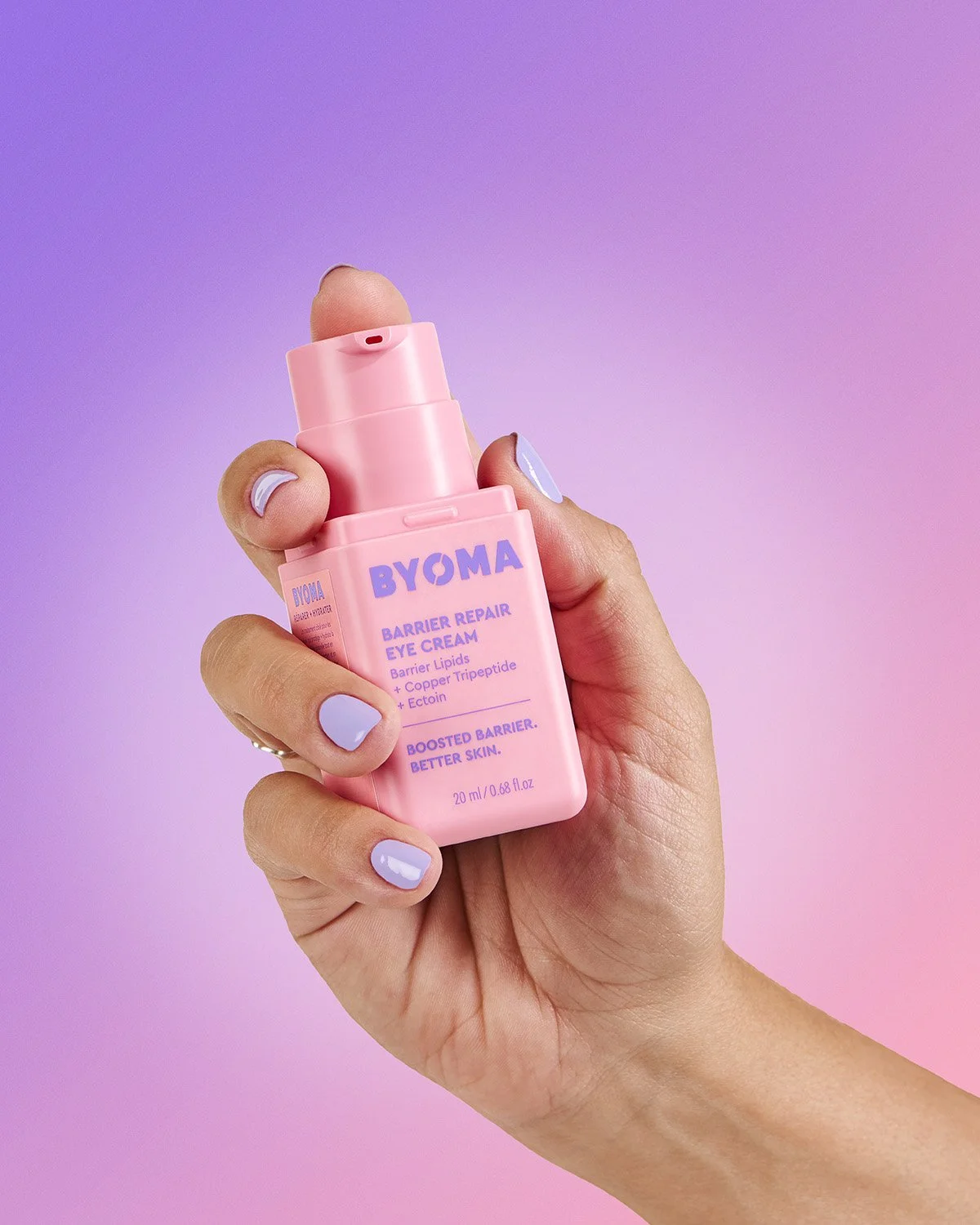Hand holding two small Byoma skincare products against a pink to purple gradient background, soft commercial product shot showing product size in context