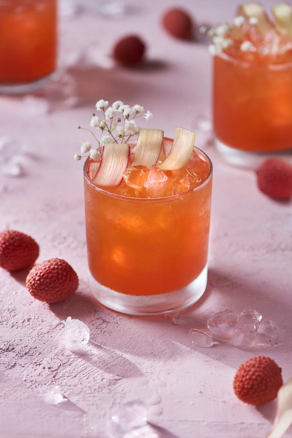 Vibrant lychee and rhubarb cocktail on the rocks garnished with fresh cranberries, shot on a soft pink surface with a light and airy feel