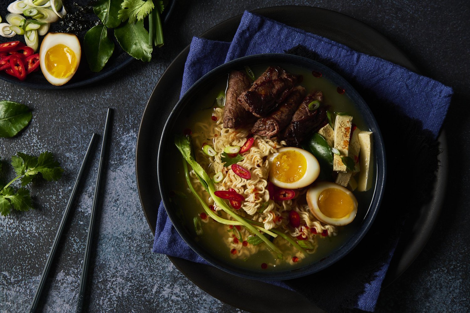  Steaming Asian noodle soup with ramen noodles, a halved soft boiled egg, sliced peppers, and fresh greens in a black bowl on a dark surface
