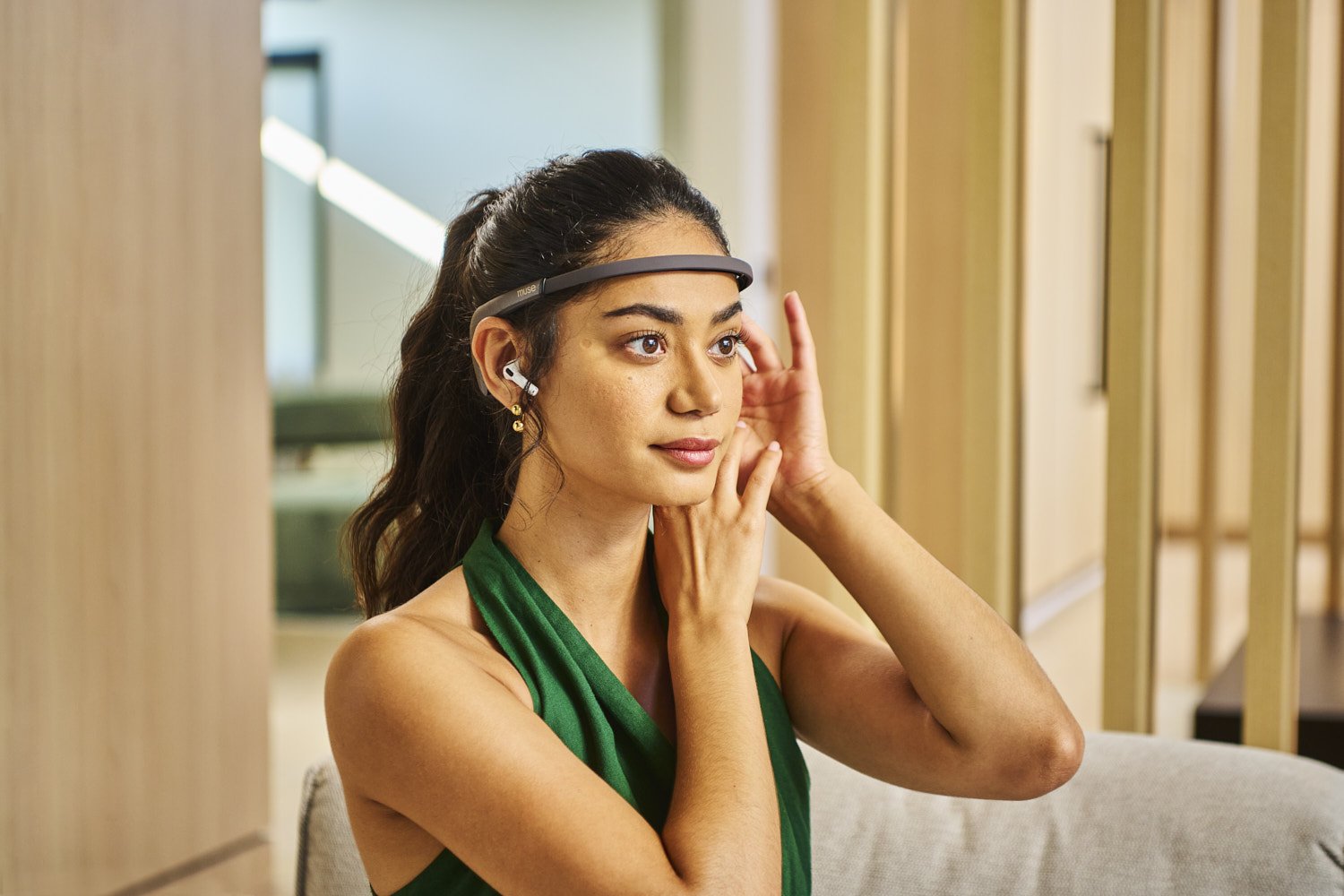 Young woman touching her face gently while wearing wireless earbuds and a sleek headband wearable device, lifestyle portrait in a bright contemporary interior setting