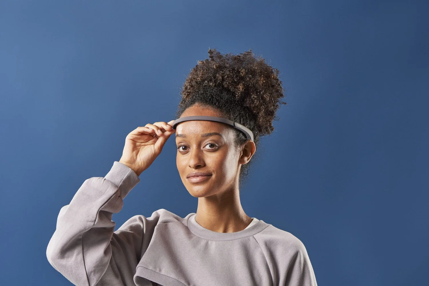 Young Black woman adjusting a tech headband wearable on her head, smiling slightly, shot against a solid blue background with clean even studio lighting