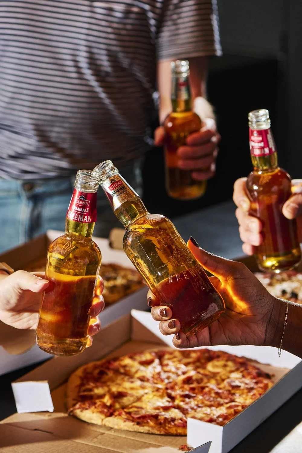 Casual gathering scene with hands reaching for beer bottles over a pizza box on a dark table, authentic lifestyle shot with warm ambient lighting