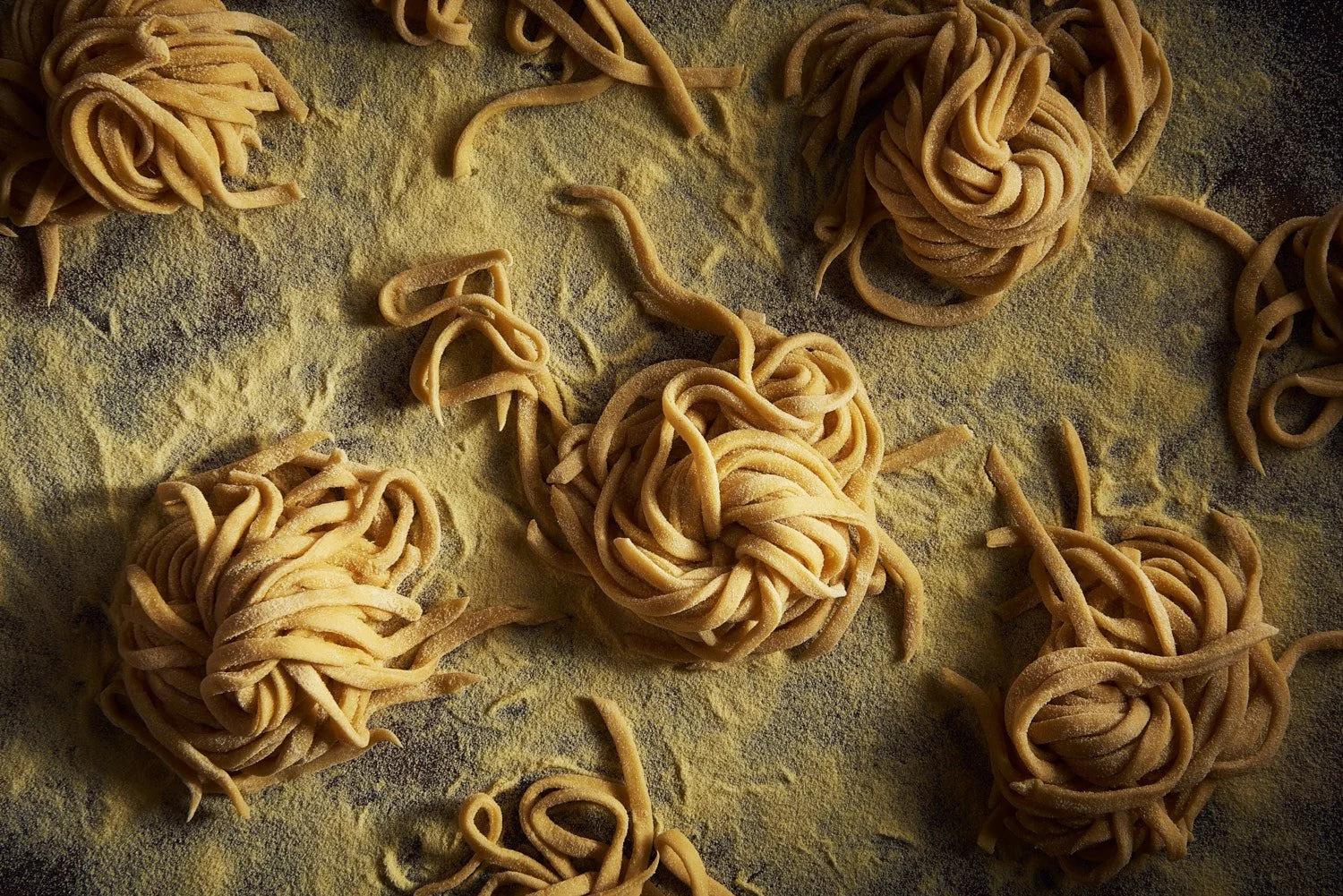 Overhead flat lay of uncooked noodle nests arranged loosely across a dark weathered surface, natural textures creating an organic and rustic food styling composition