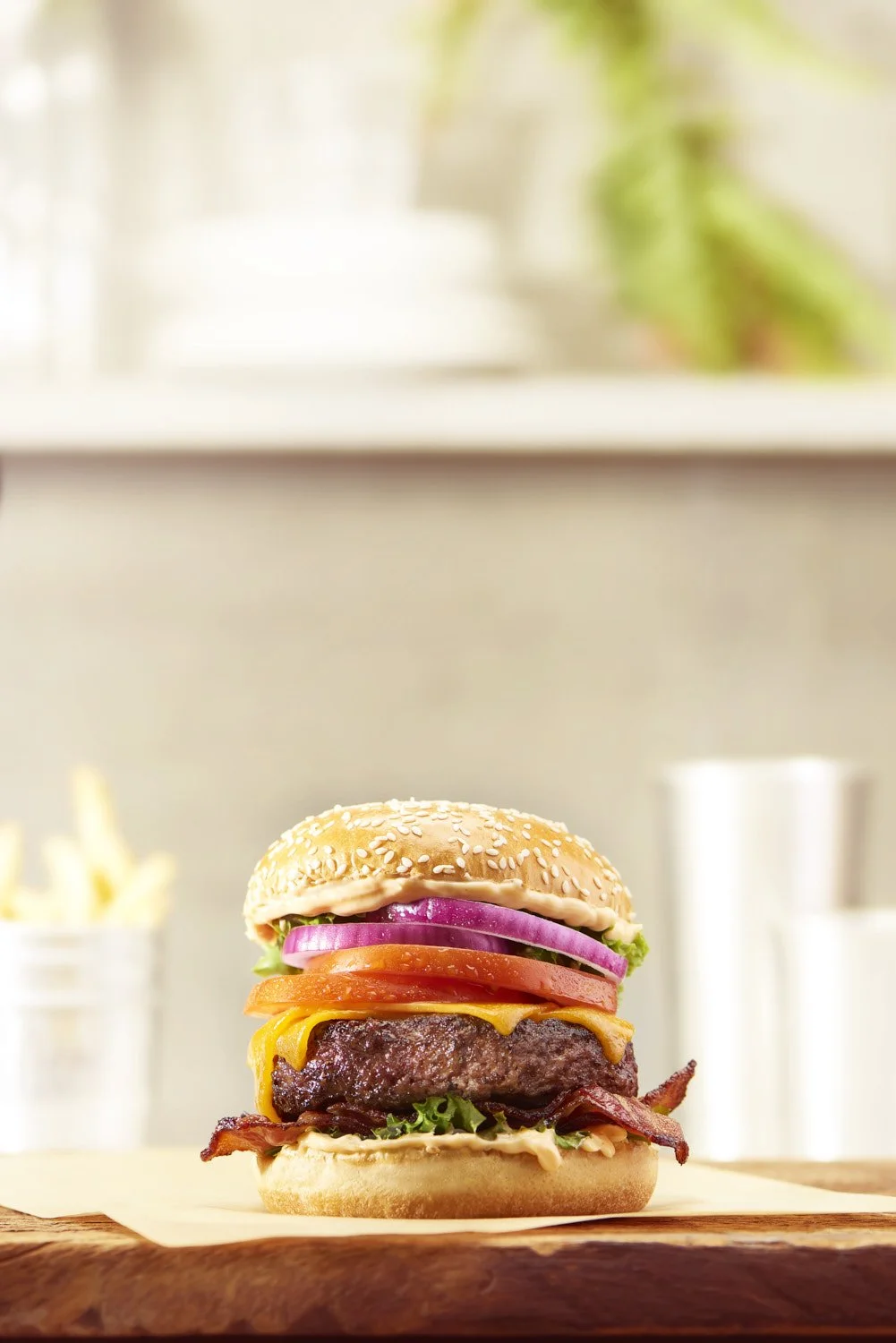 Stacked gourmet burger with lettuce, tomato, red onion, and condiments on a sesame bun, photographed in a bright airy kitchen setting with soft natural light