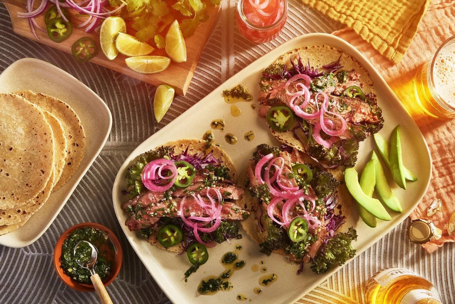 Overhead styled taco shot featuring open tortillas with sliced steak, floral garnishes, avocado, and sides, arranged artfully on a warm neutral background
