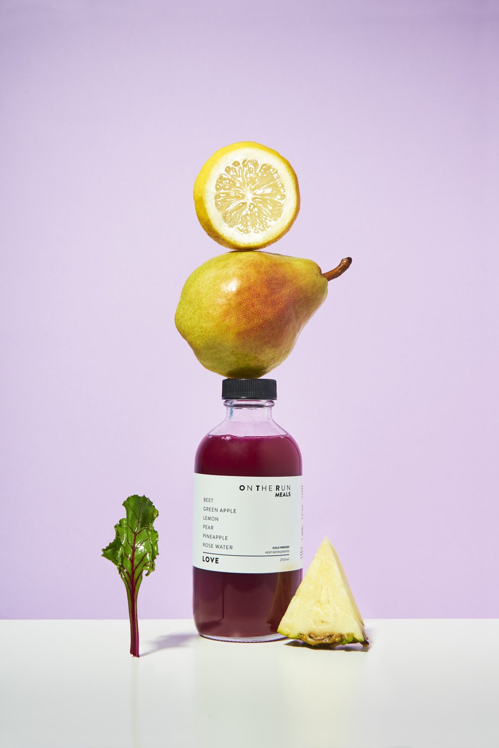 Playful stacked composition of a lemon slice balanced on a pear balanced on a small beverage bottle, shot on a clean lavender purple background
