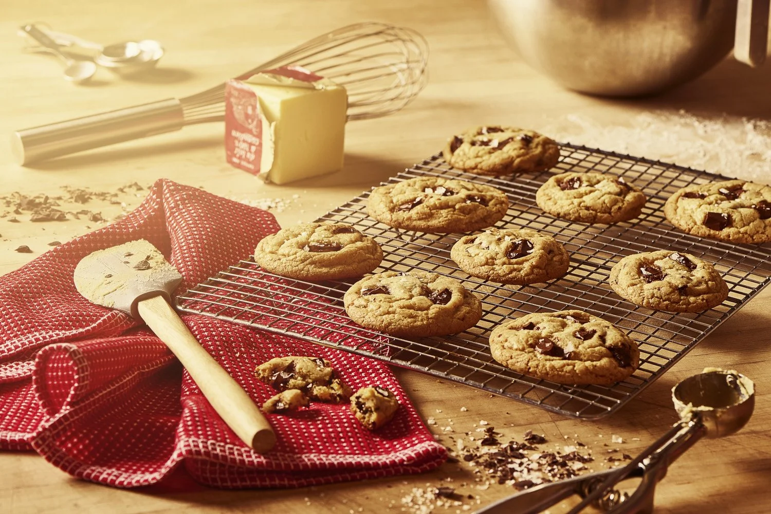 Freshly baked chocolate chip cookies arranged on a wire rack beside a red linen cloth and a rolling pin, shot with warm golden side lighting