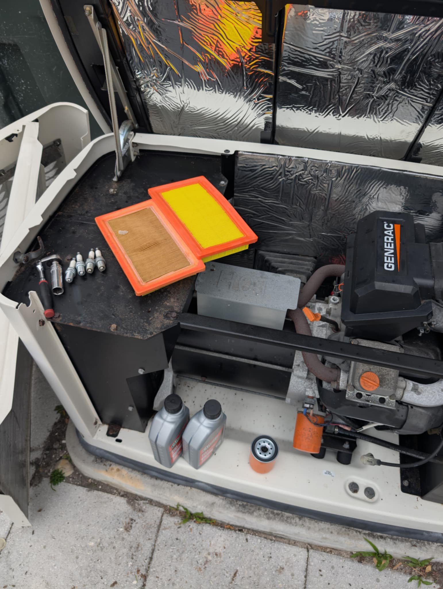 View of the engine compartment of a small recreational vehicle or golf cart, with tools, oil bottles, air filters, and engine components visible, and a reflective heat shield or insulation in the background.