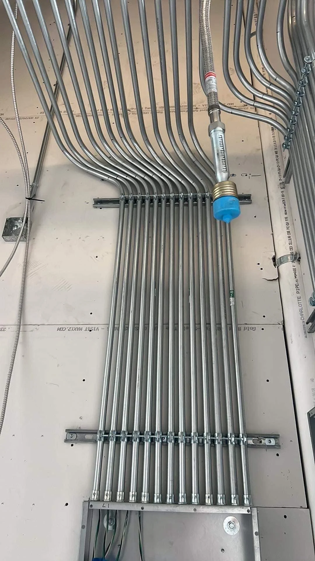 Metal piping and tubing installed on a wall, with a pressure gauge and a blue cap, in a building under construction or maintenance.