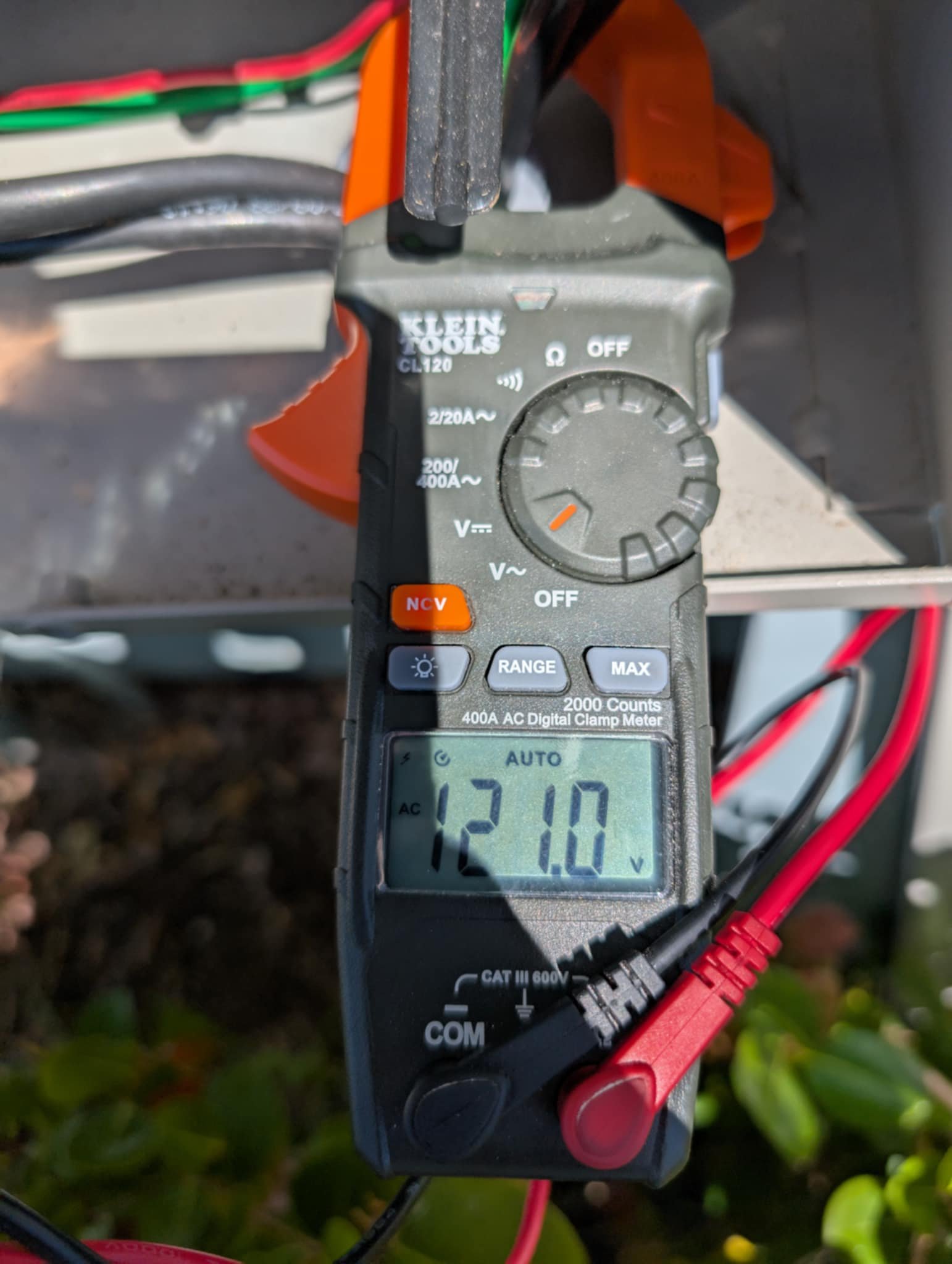 Digital clamp meter measuring 12.10 volts in an outdoor setting, with red and black test leads connected to electrical wires.