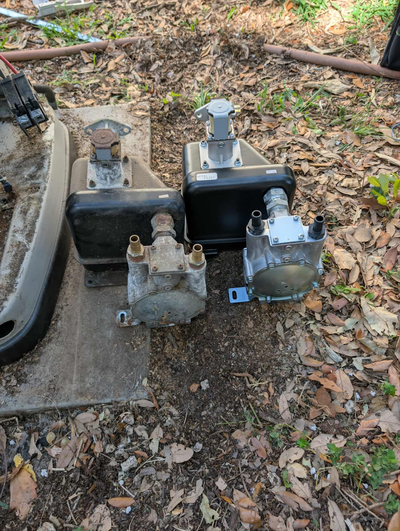 Several mechanical parts and components on the ground outdoors, including a black rectangular box, a metallic motor with wires, and a rusty, weathered component, surrounded by dirt, leaves, and grass.