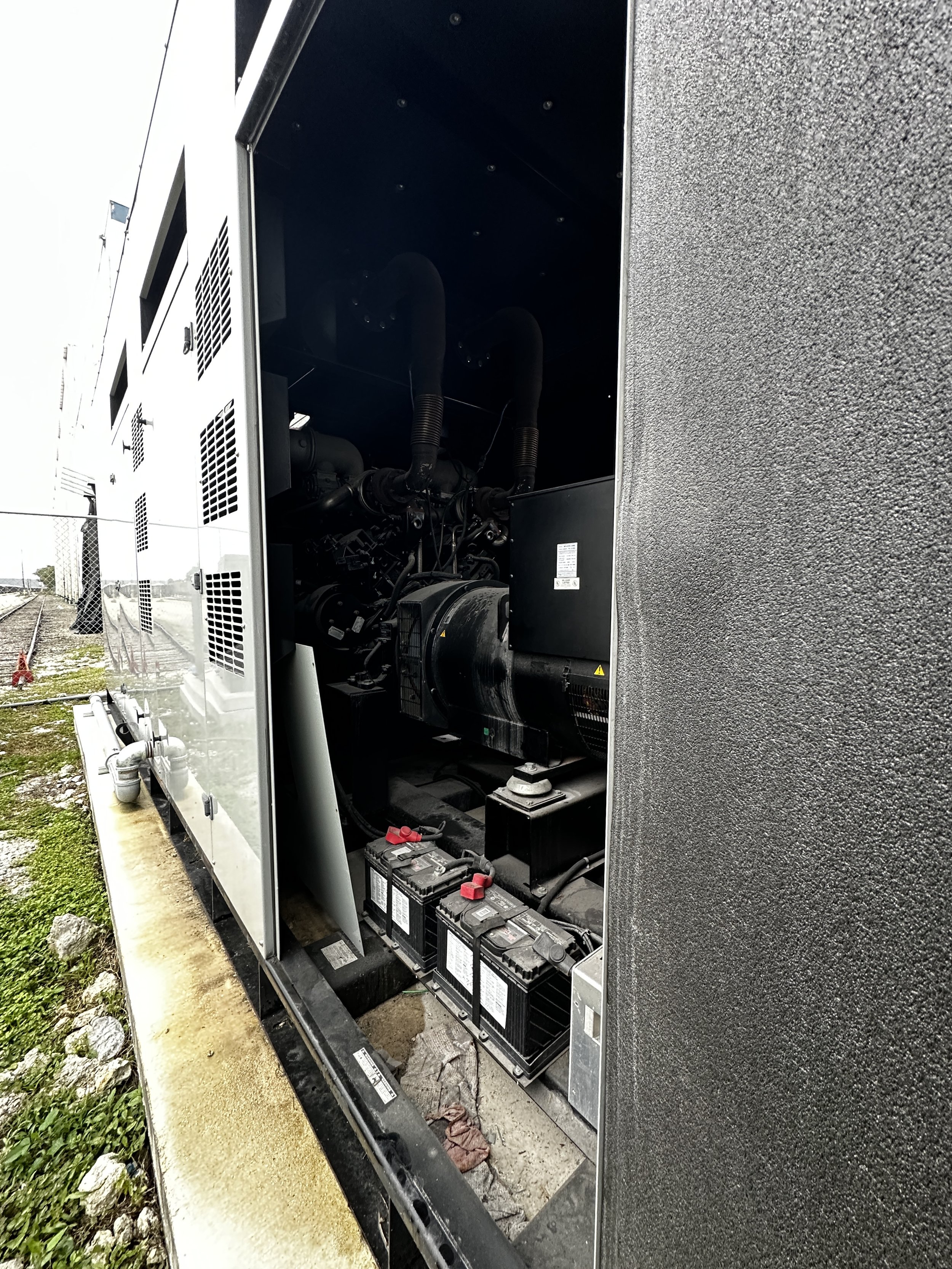 Exterior view of a generator or industrial equipment with open panels showing internal components, located outdoors near train tracks.