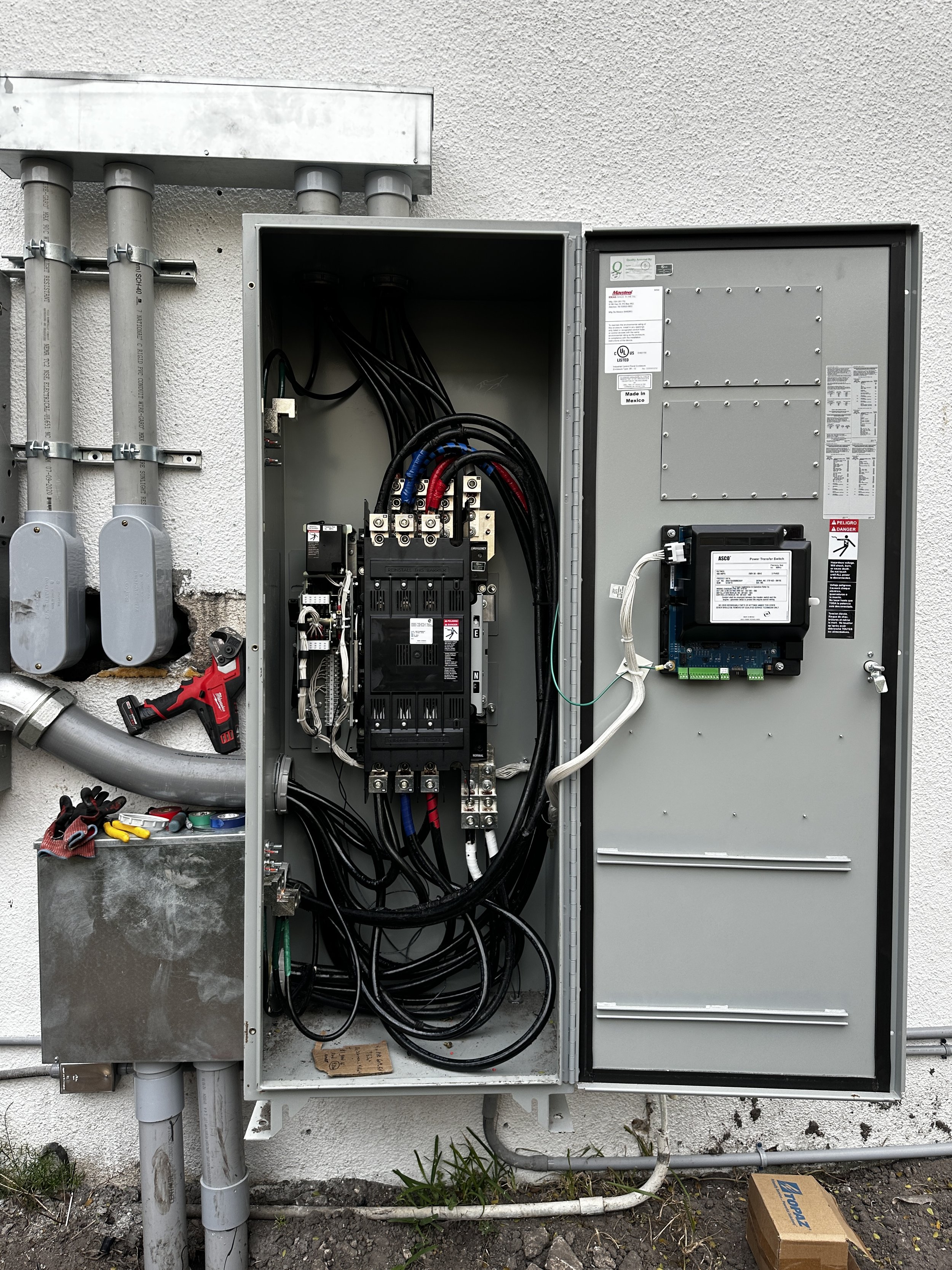 Open electrical control panel with wires, circuit breakers, and electronic components mounted in a gray metal box on an exterior wall. Pliers, tools, and a drill are nearby.