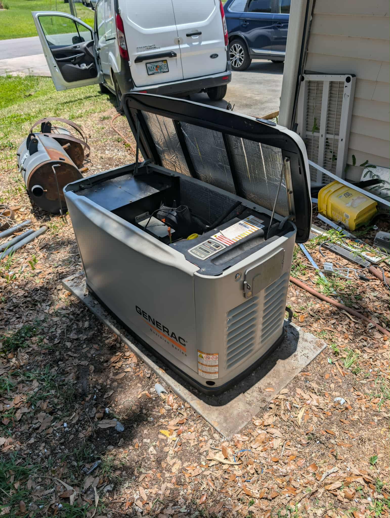 An open generator outdoors on a concrete pad surrounded by leaves, with a white van and blue car in the background.