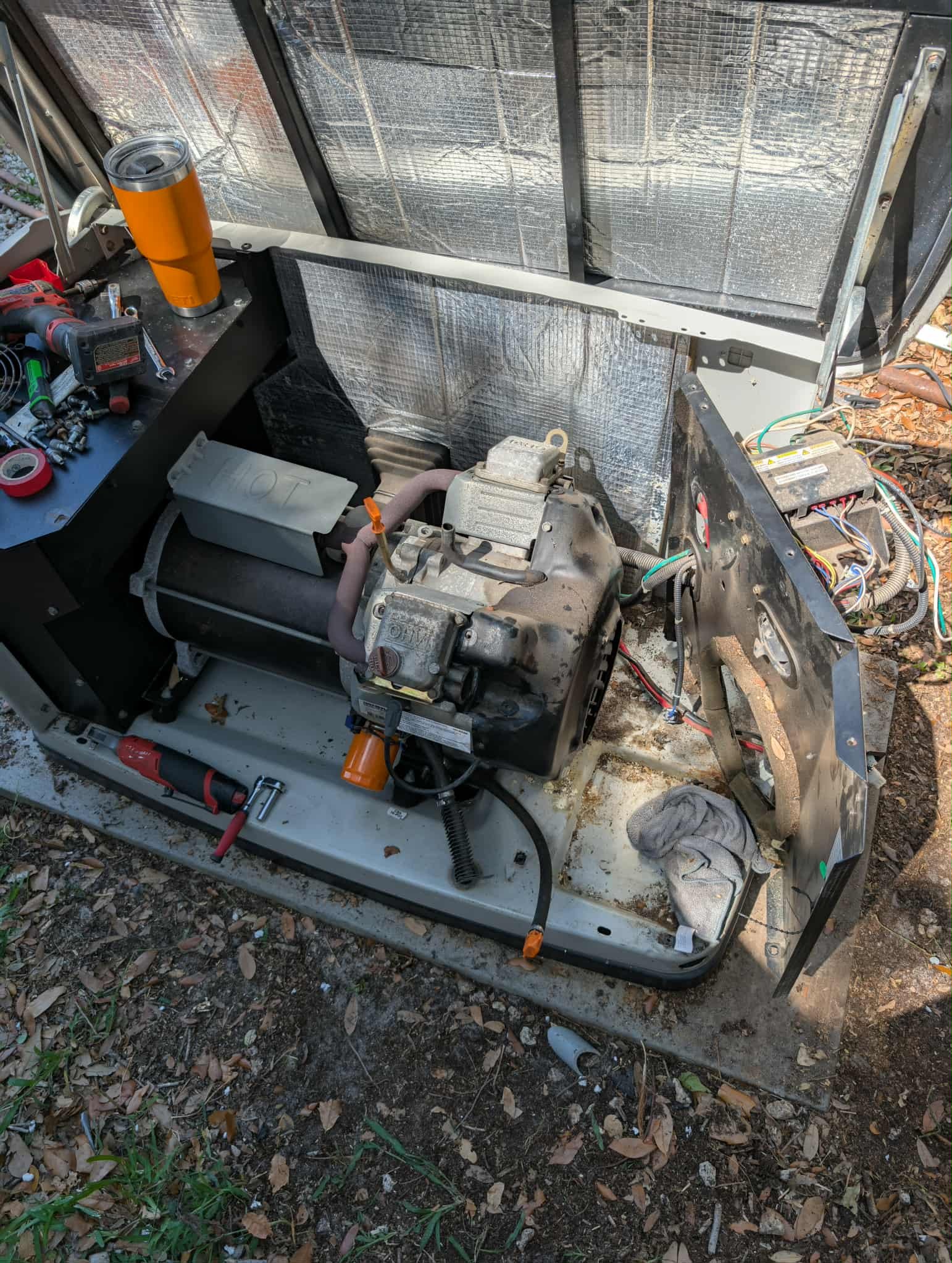 Partial view of mechanical outdoor equipment with removed panels, showing an engine, wires, tools, and a towel, on a dirty surface surrounded by leaves.