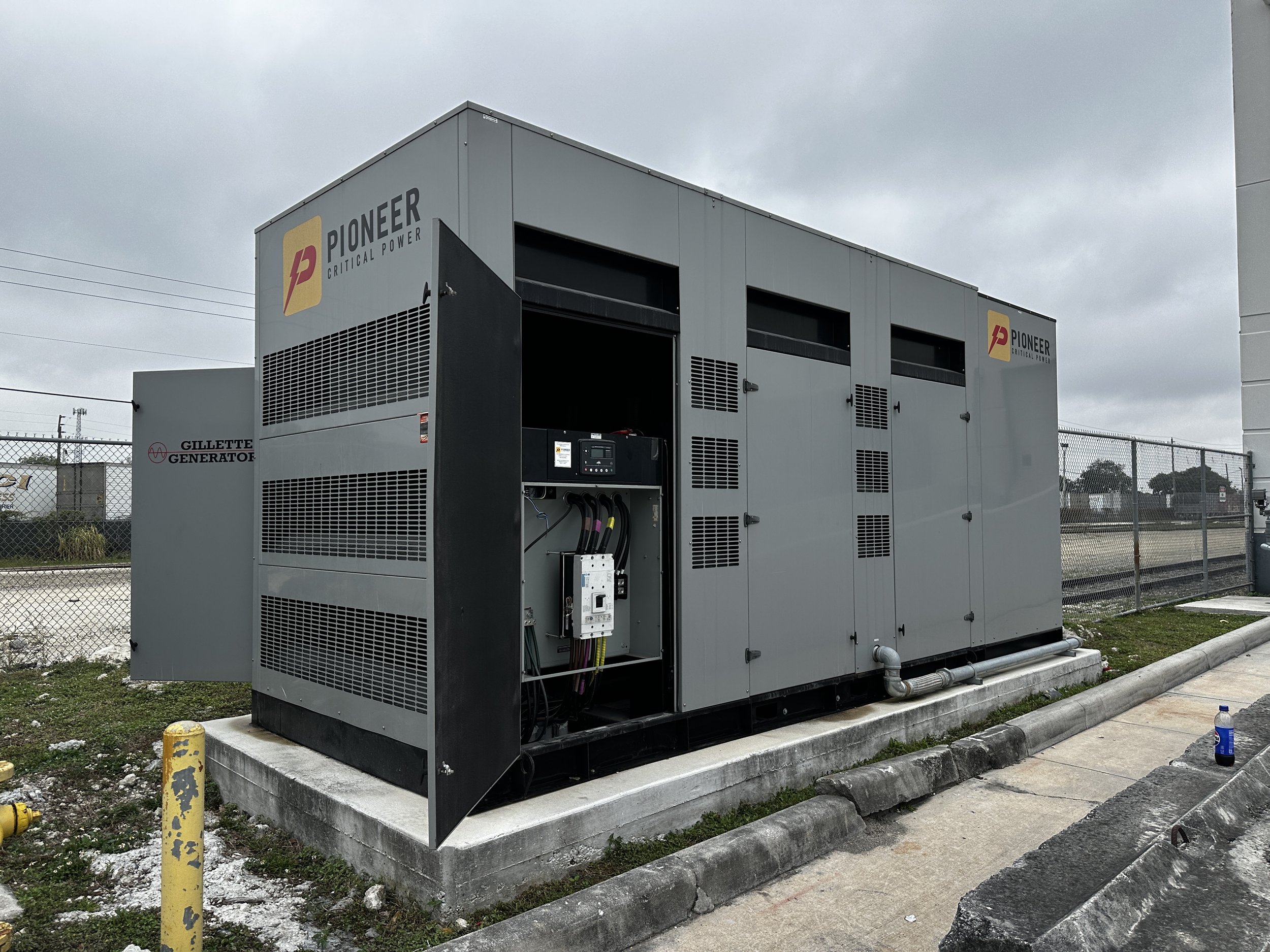 Large industrial generator outdoors next to a paved sidewalk, fenced area in background, overcast sky.