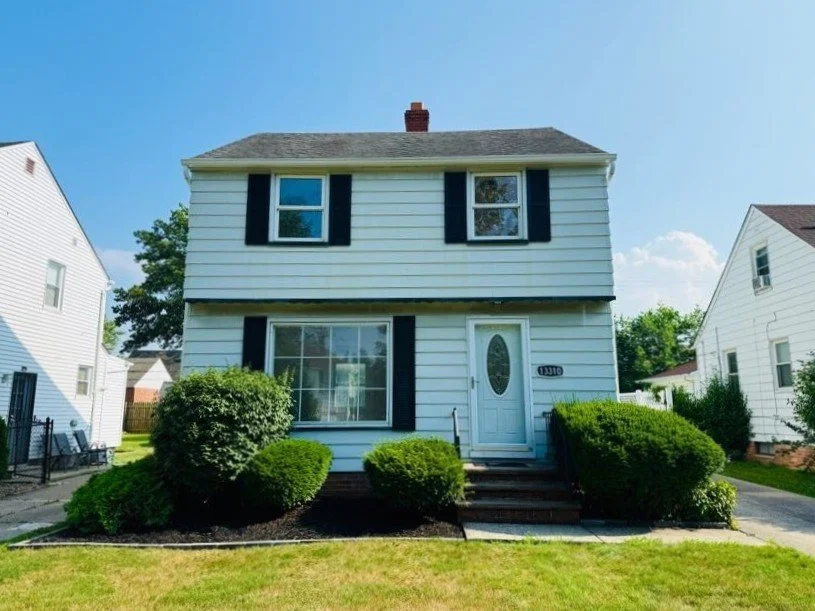 Front view of a two-story white house with black shutters, a white door, and neatly trimmed bushes in the front yard. Clear blue sky in the background.