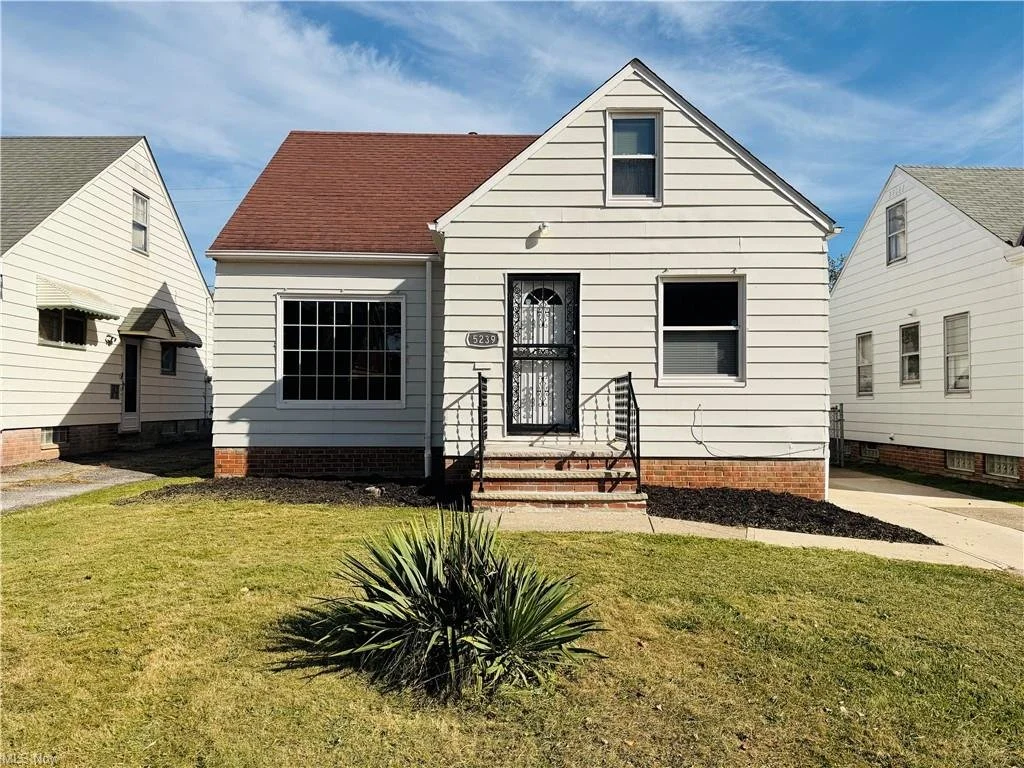 A small white house with a red roof and front steps, surrounded by a well-kept lawn and neighboring houses, under a blue sky.
