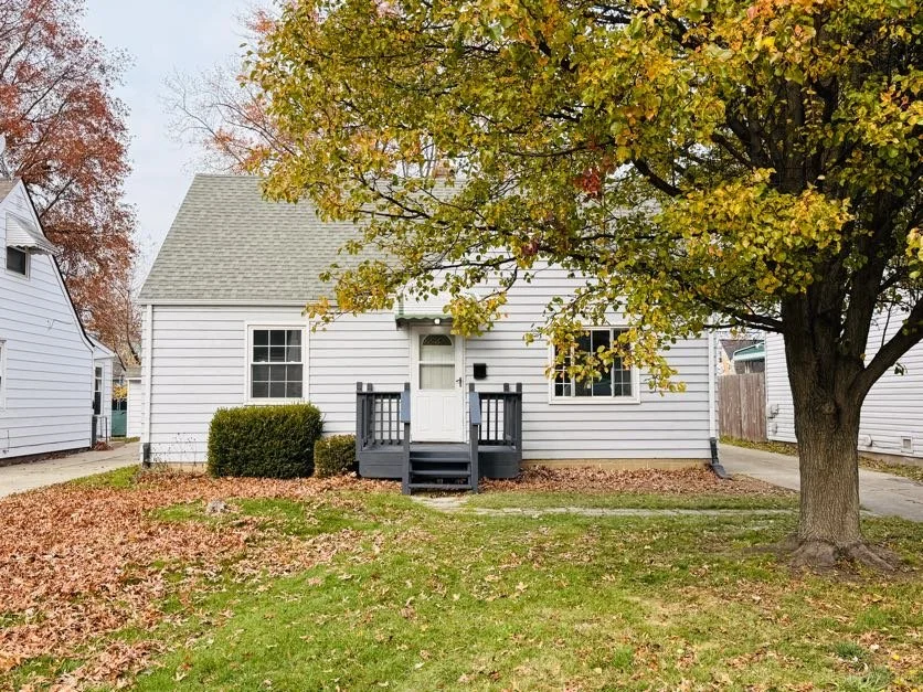 A small white house with a gray roof, a front door with small stairs, and two windows. There is a large leafy tree in the front yard and fallen leaves on the grass.