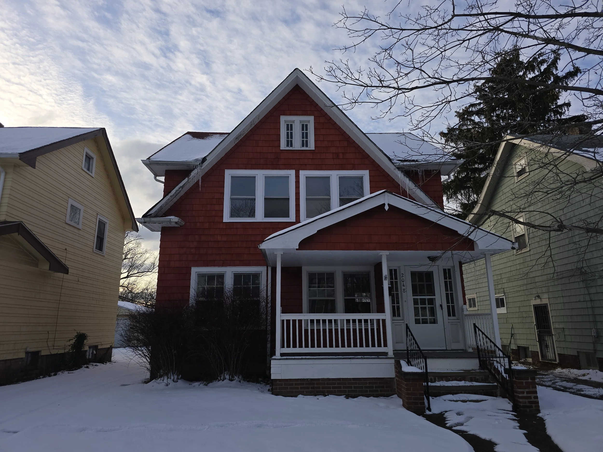 Red three-story house with white trim, covered porch, brick steps, surrounded by snow in winter, with neighboring yellow and green houses on either side, and a cloudy sky above.