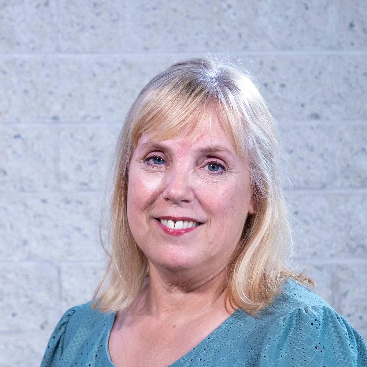 Woman with blonde hair and blue top standing against a light brick wall.