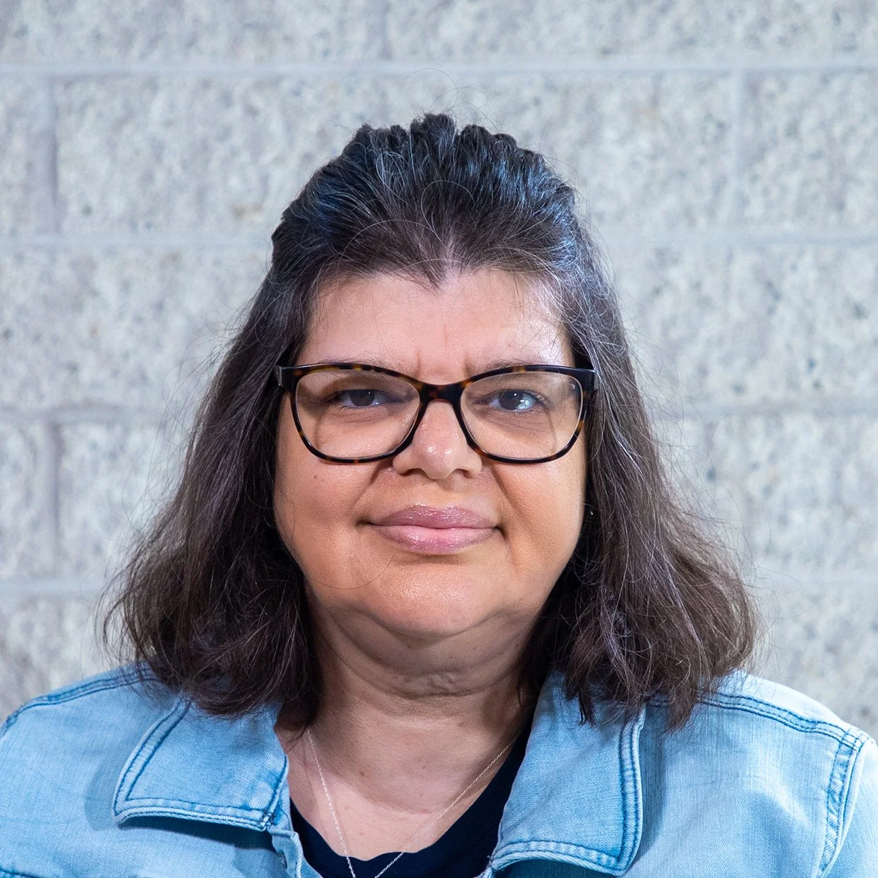 Woman with brown hair and glasses wearing a blue shirt standing in front of a gray brick wall.