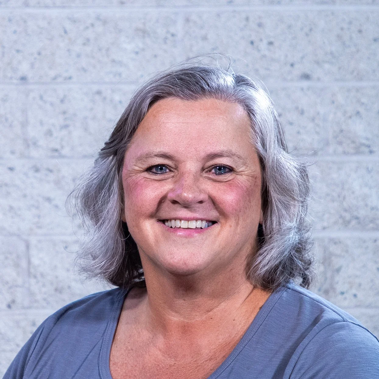 Woman with gray hair smiling in front of a brick wall, wearing a blue top and necklace.