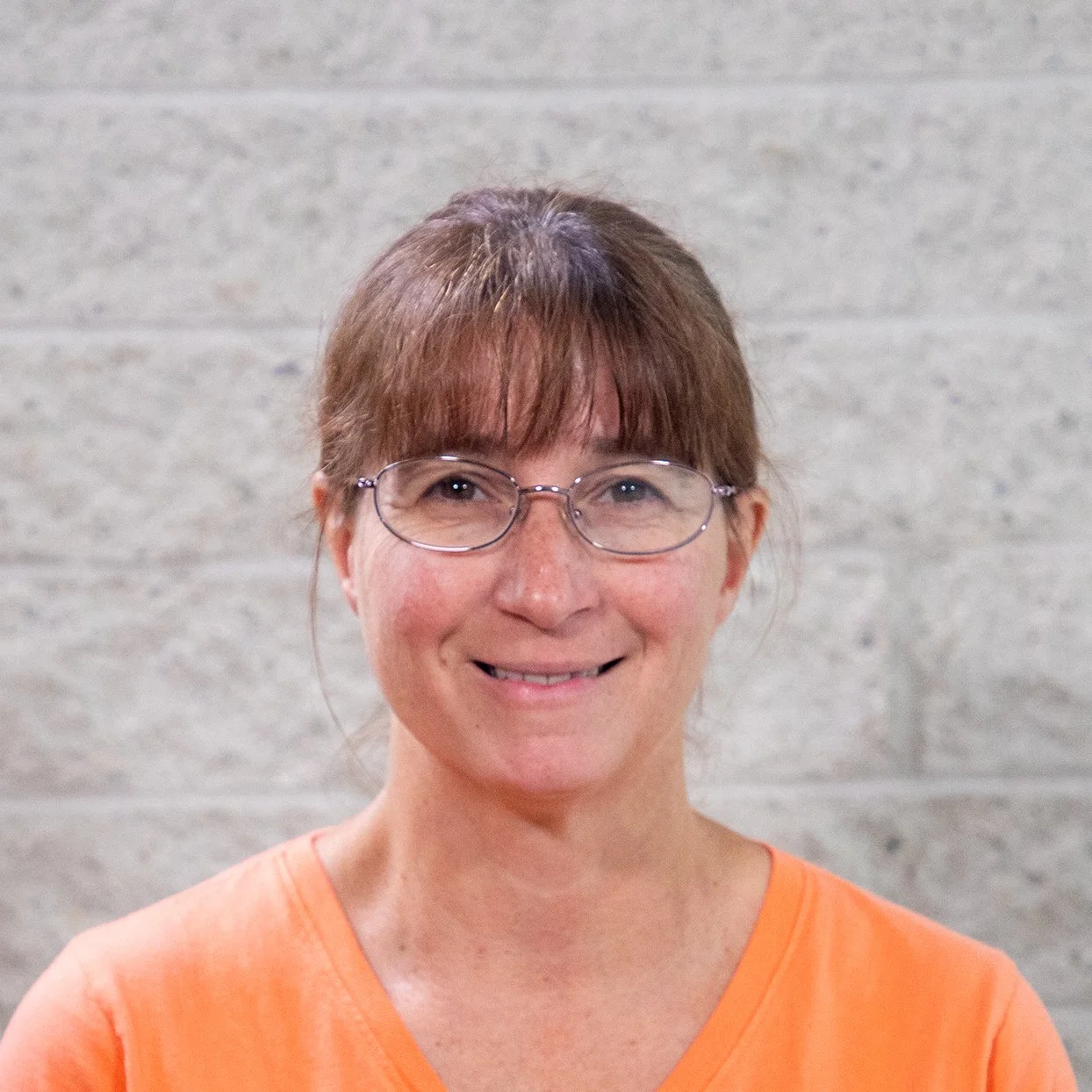 Woman with glasses and a purple shirt smiling in front of a white brick wall.