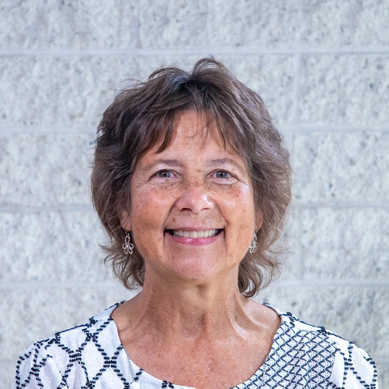 Smiling woman with short brown hair in a colorful shirt against a light brick wall.