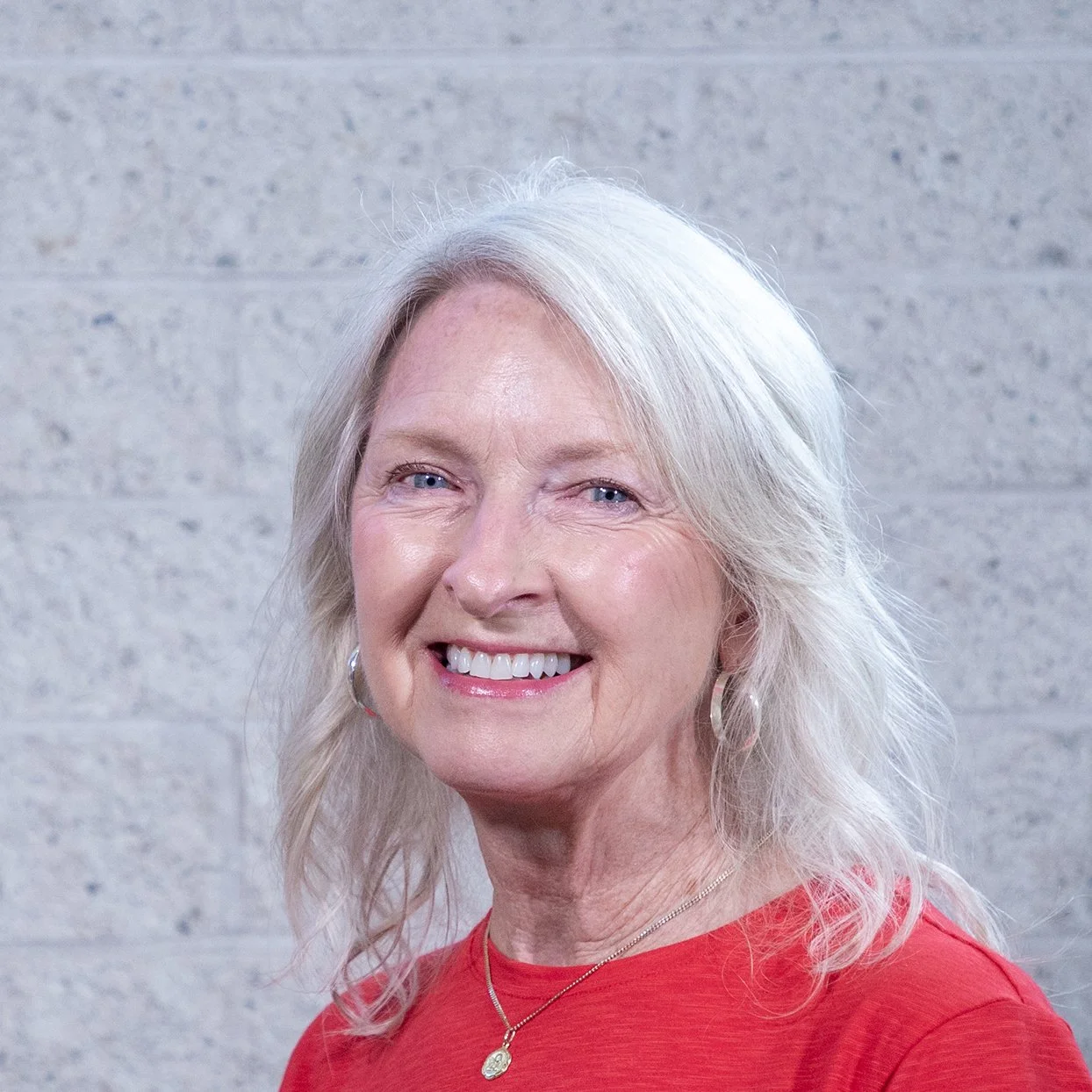 Smiling woman with long white hair standing against a stone wall.