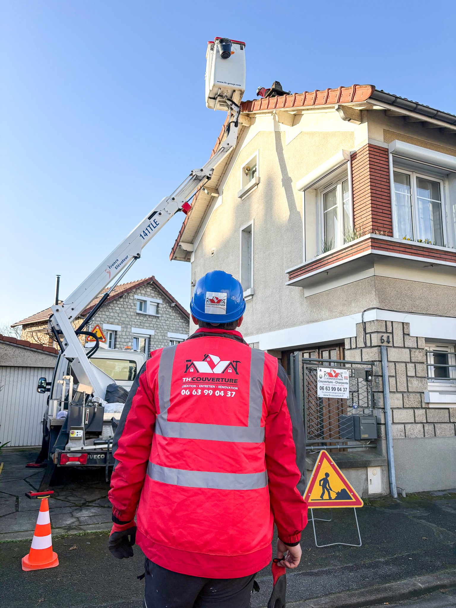 Un ouvrier en veste rouge et casque bleu observe une plateforme élévatrice près d'une maison en construction ou en rénovation.