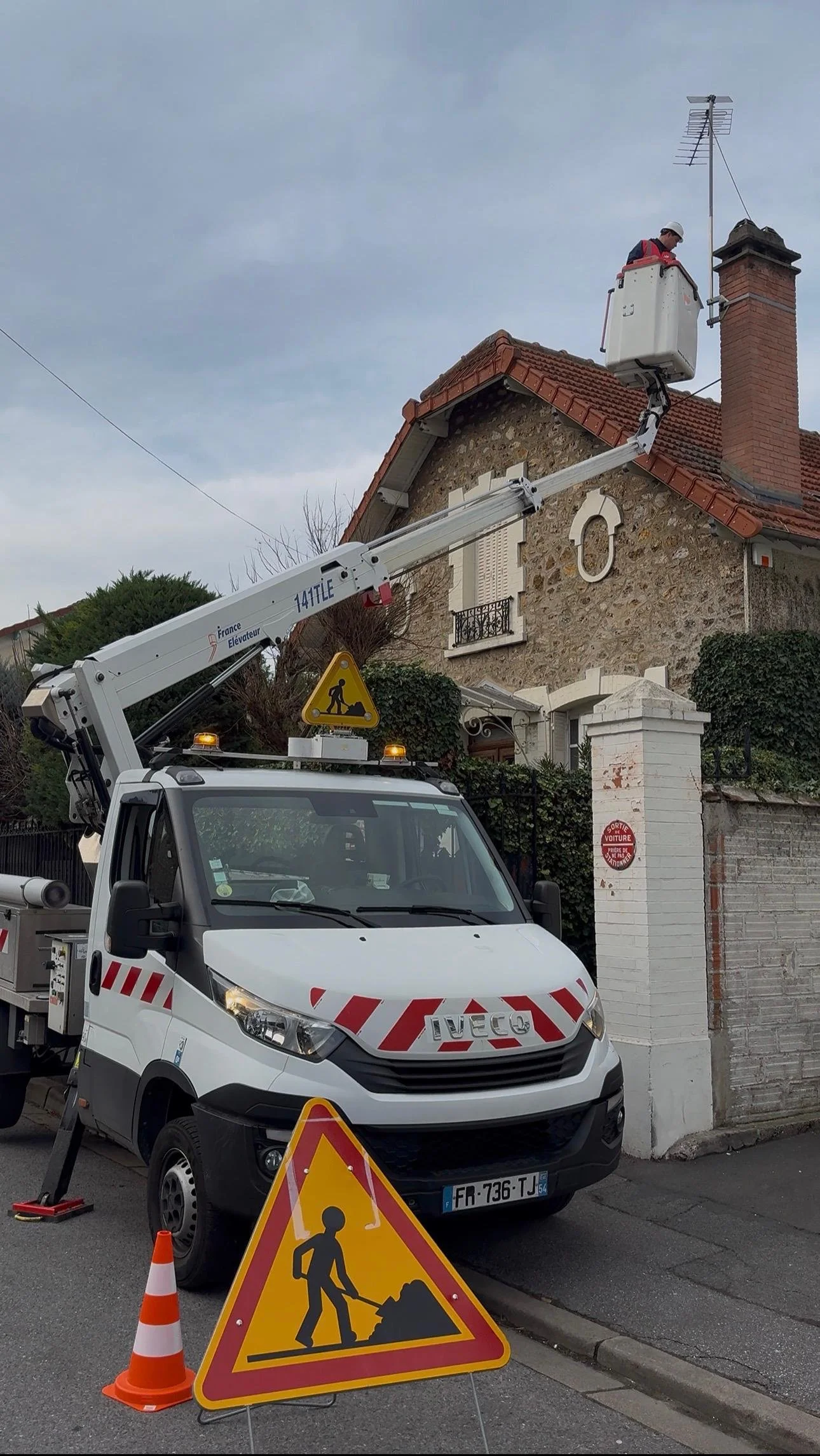 Une voiture de service avec un équipement de levage en position à côté d'une maison, un ouvrier en train de travailler en hauteur dans le SUV.